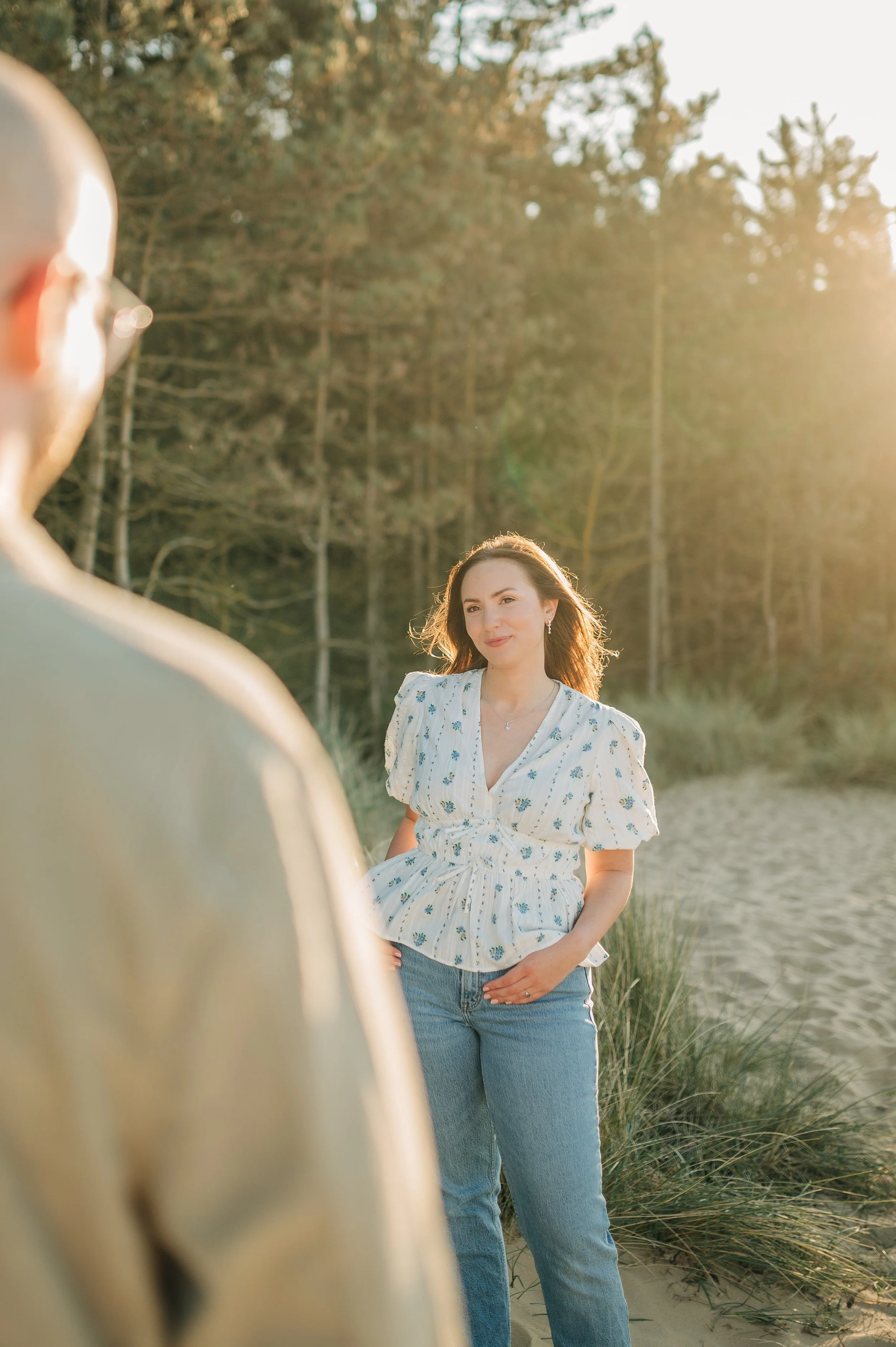 Norfolk Wedding Photographer.  A woman standing outdoors on a beach during sunset, wearing a white blouse with blue floral patterns and blue jeans, smiling and looking at a person who is partially visible in the foreground.
