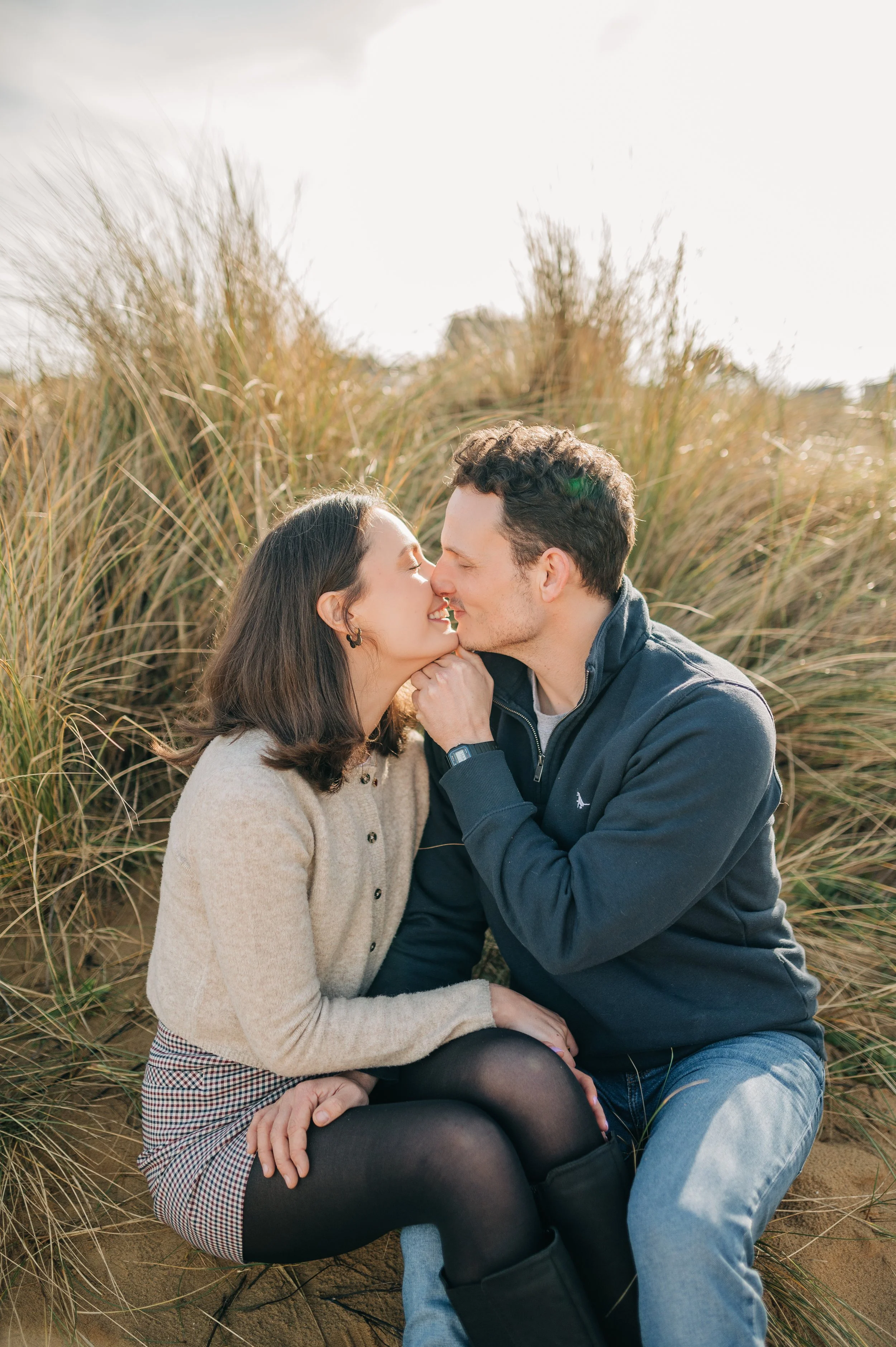 Norfolk Wedding Photographer.  A couple sitting on the ground amid tall grass, about to kiss outdoors on a sunny day.
