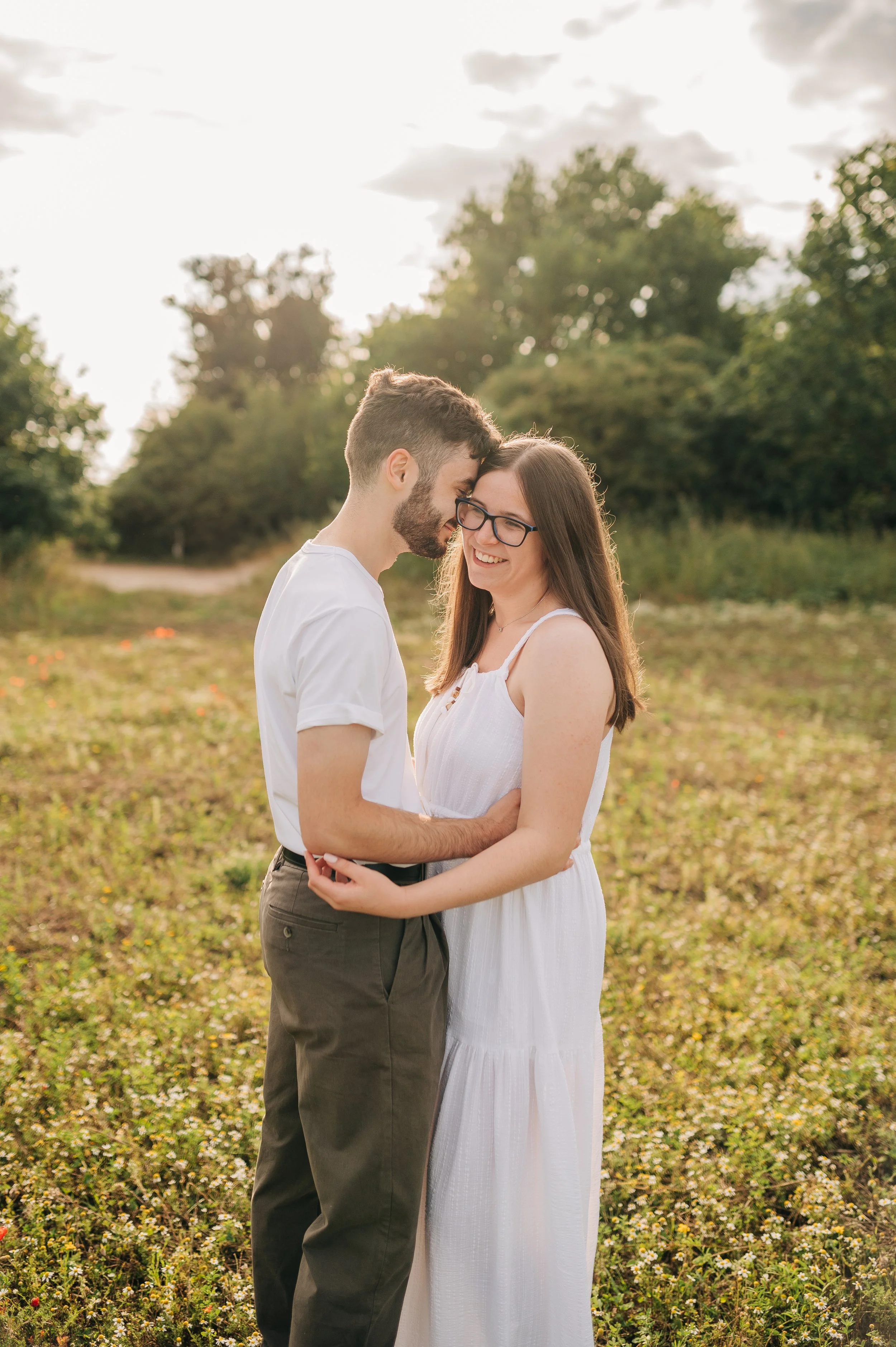 Norfolk Wedding Photographer.  A young couple stands close together in a field, smiling, with sunlit trees and cloudy sky in the background.