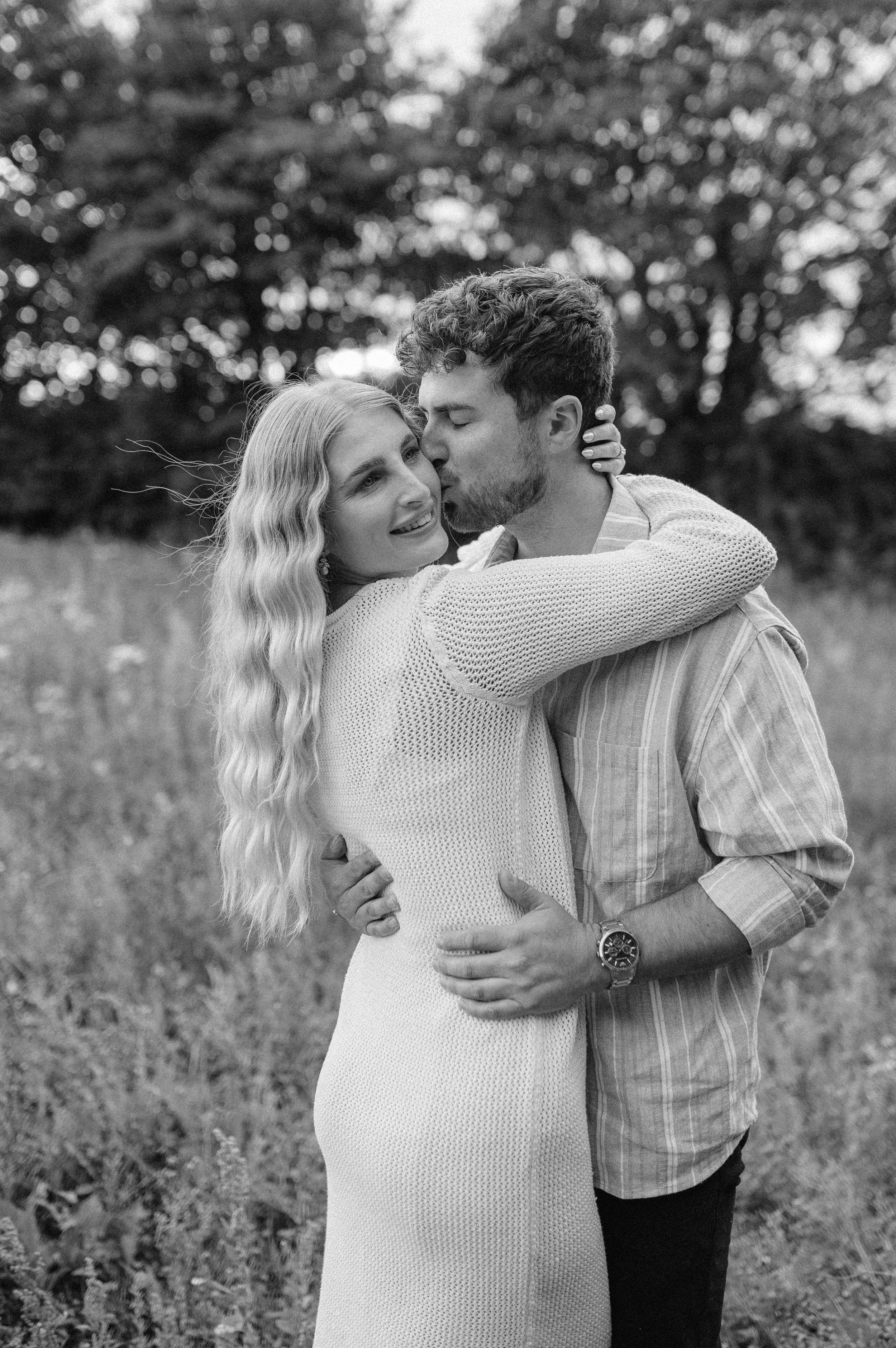Norfolk Wedding Photographer.  A black and white photo of a couple embracing outdoors, with the man kissing the woman on the cheek while she smiles, in a grassy field with trees in the background.