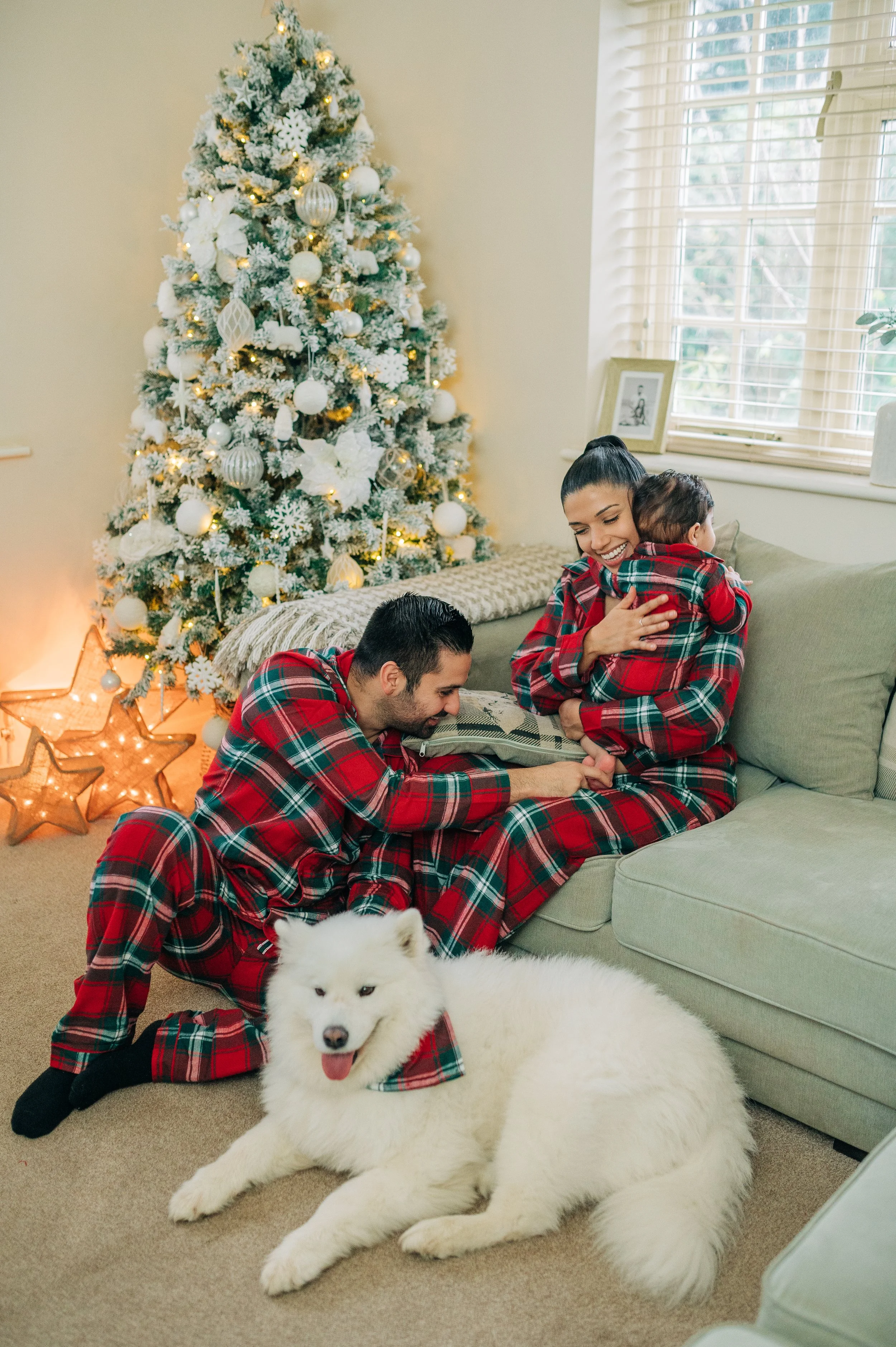 Norfolk Family Photographer.  A family celebrates Christmas in matching red plaid pajamas in a decorated living room with a Christmas tree, a dog, and holiday lights.