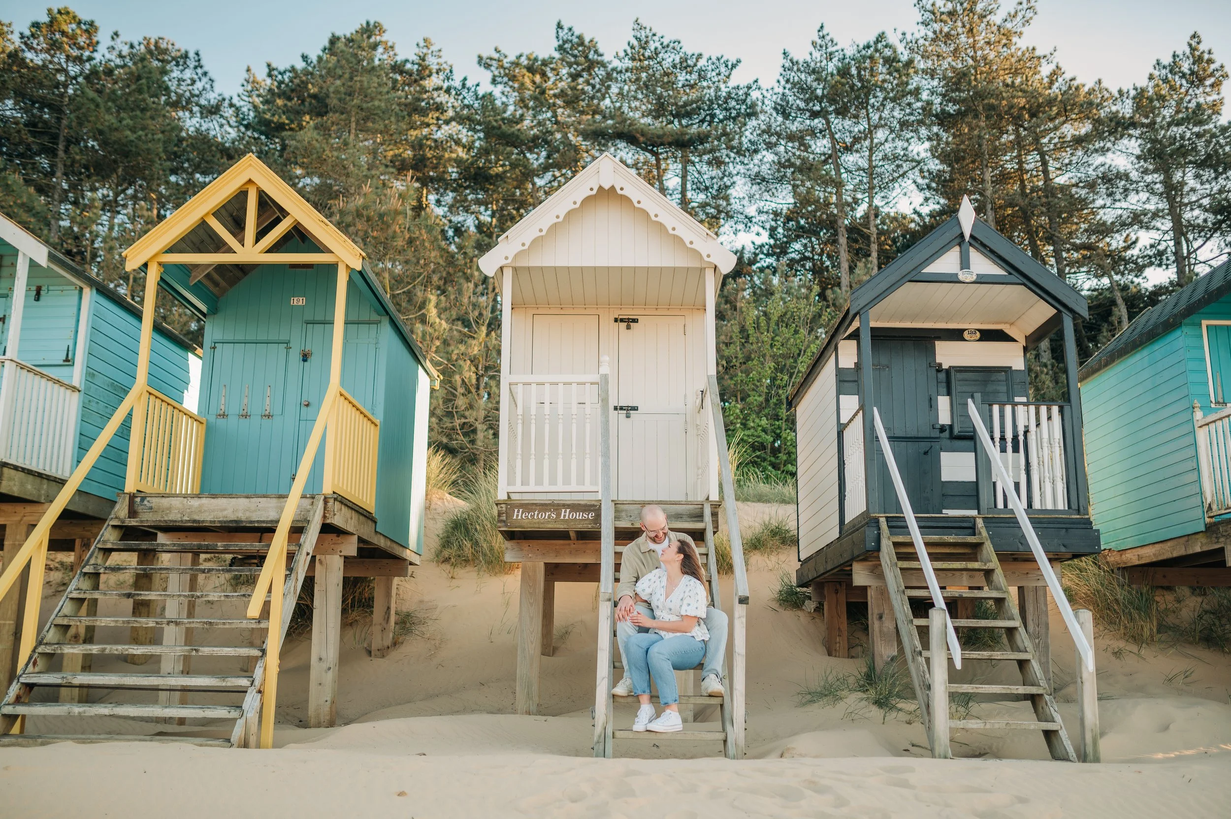 Norfolk Wedding Photographer.  A couple sitting on steps in front of colorful beach huts on a sandy beach, with trees in the background.