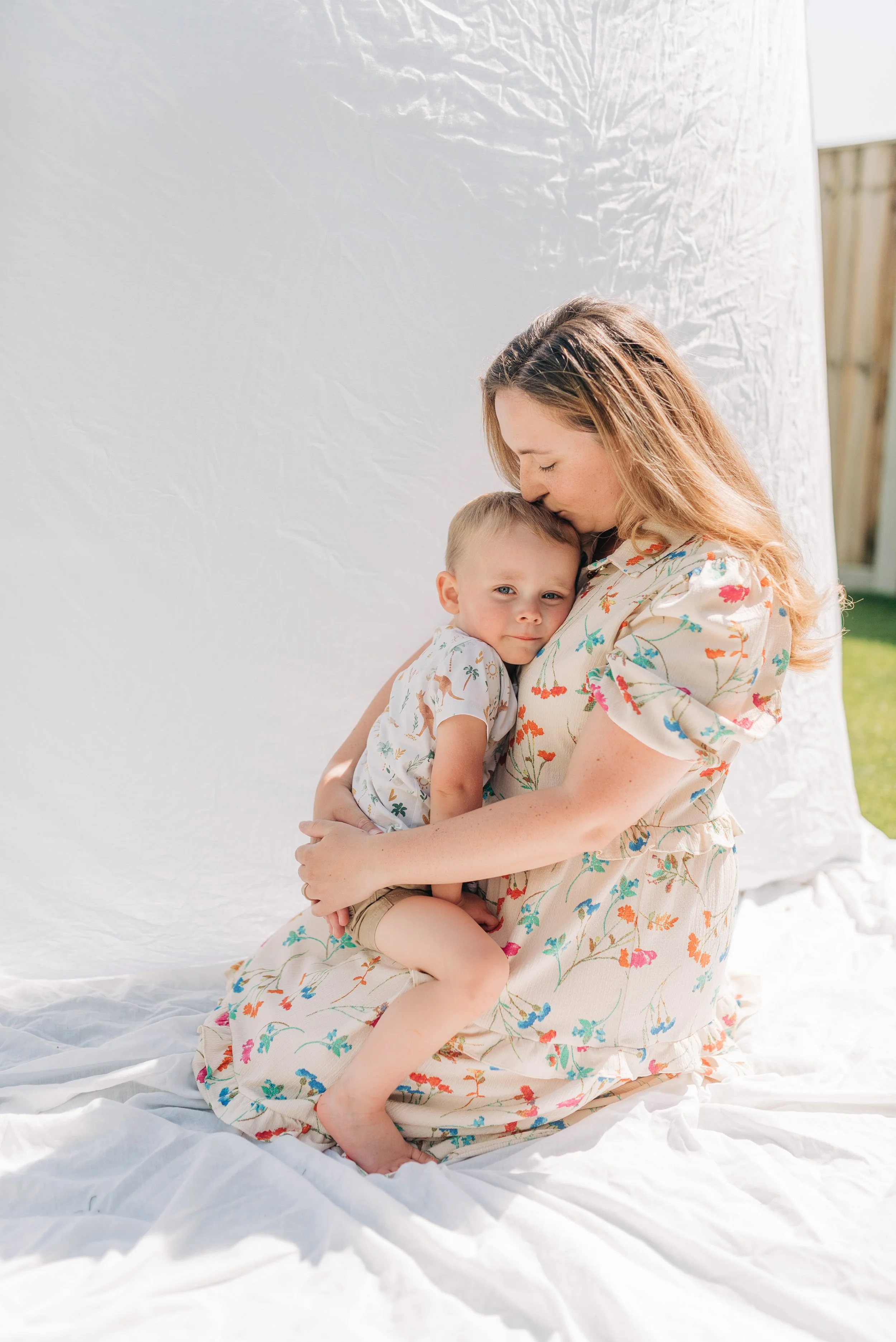 Norfolk Family Photographer.  A mother and young boy share a hug outdoors on a sunny day, with the mother gently kissing the boy's head.