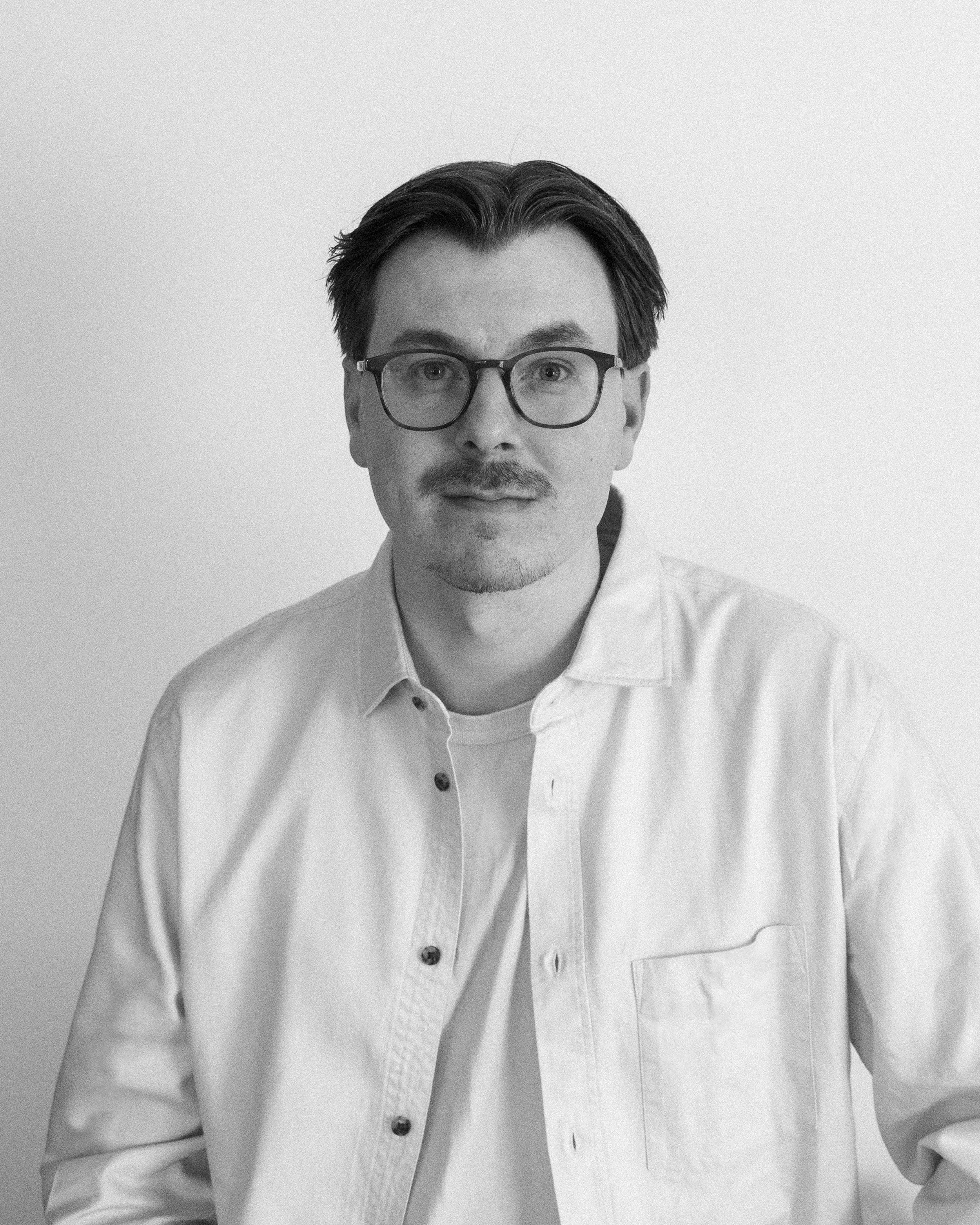 Black and white photo of a man with glasses, short dark hair, a mustache, and goatee, wearing a button-up shirt, against a plain light background.