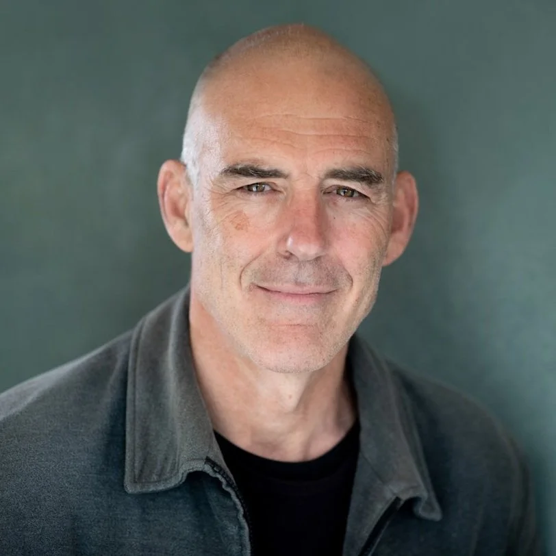 Close-up portrait of a middle-aged man with a shaved head, slight smile, wearing a dark gray collared shirt, against a plain background.