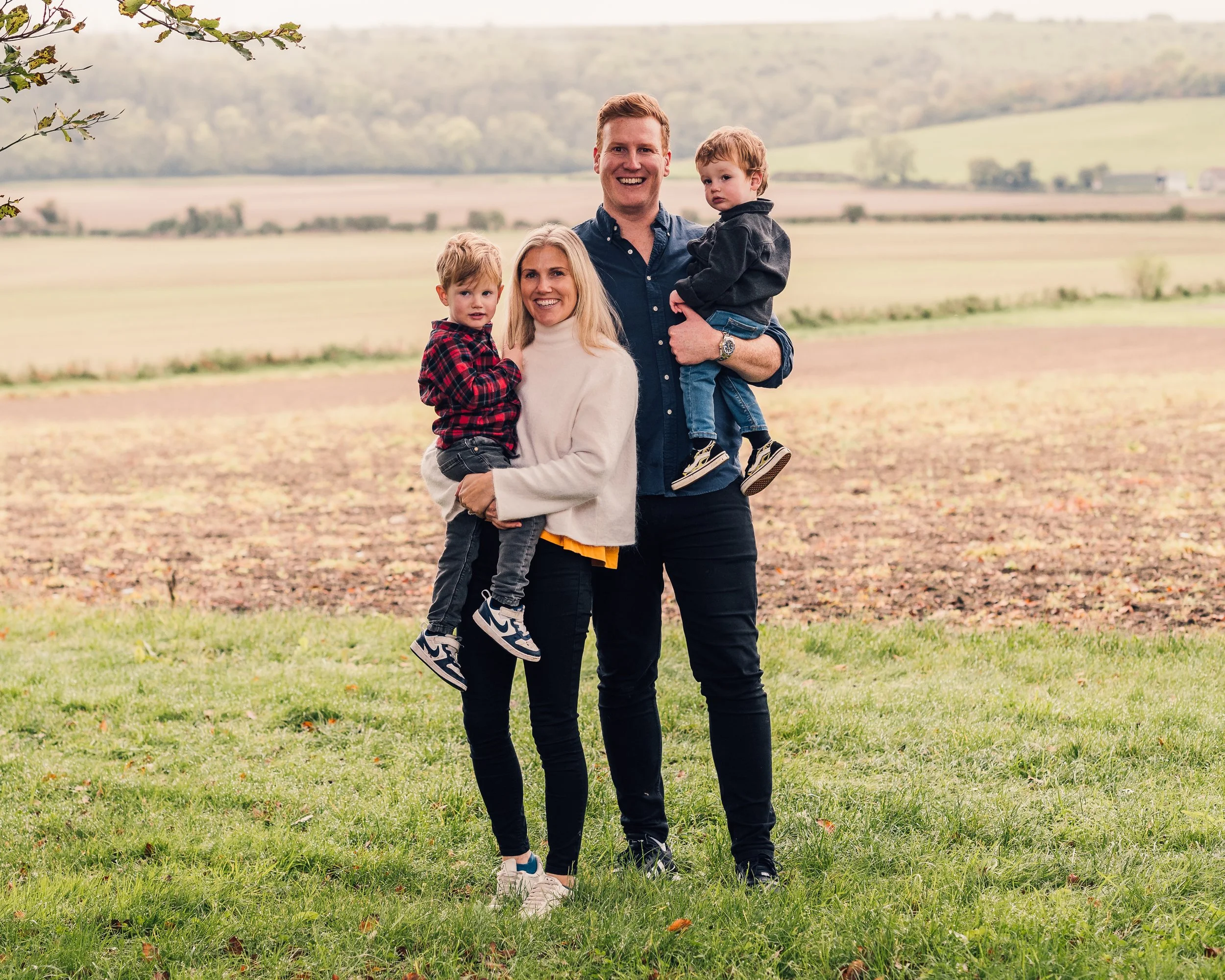 A family of four standing outdoors in a field with rolling hills in the background, smiling and posing for a photo.