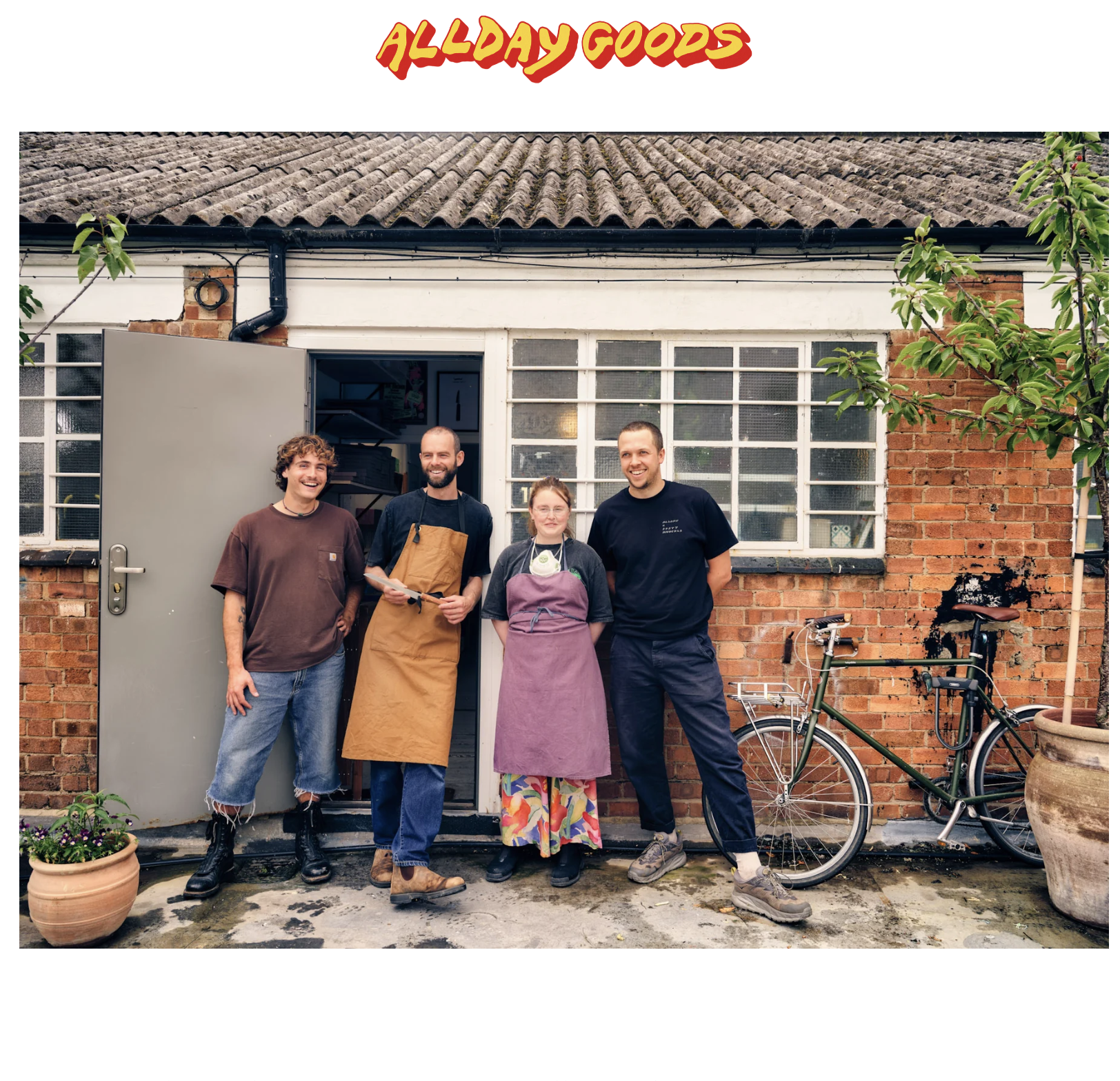 Four people stand outside a brick building with large windows, smiling. One person is holding a clipboard, and two are wearing aprons. A bicycle is parked beside them, and potted plants are nearby. The top of the image features text that says 'ALlDAY GOODS' in bold, colorful letters.