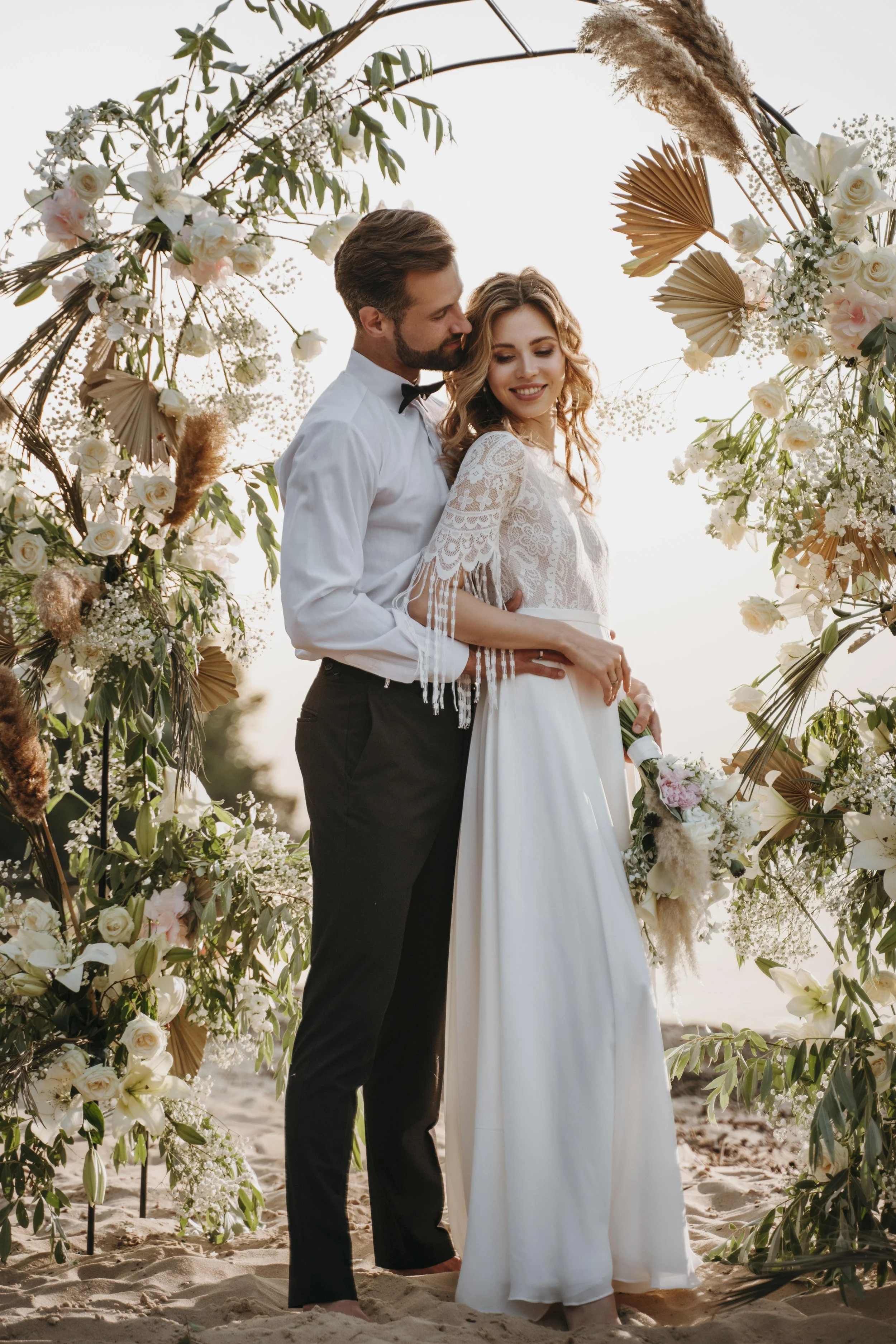 Un couple en mariée et marié sur la plage, entourés d'un cercle floral de fleurs blanches et de feuillage, lors d'une cérémonie nuptiale en plein air.