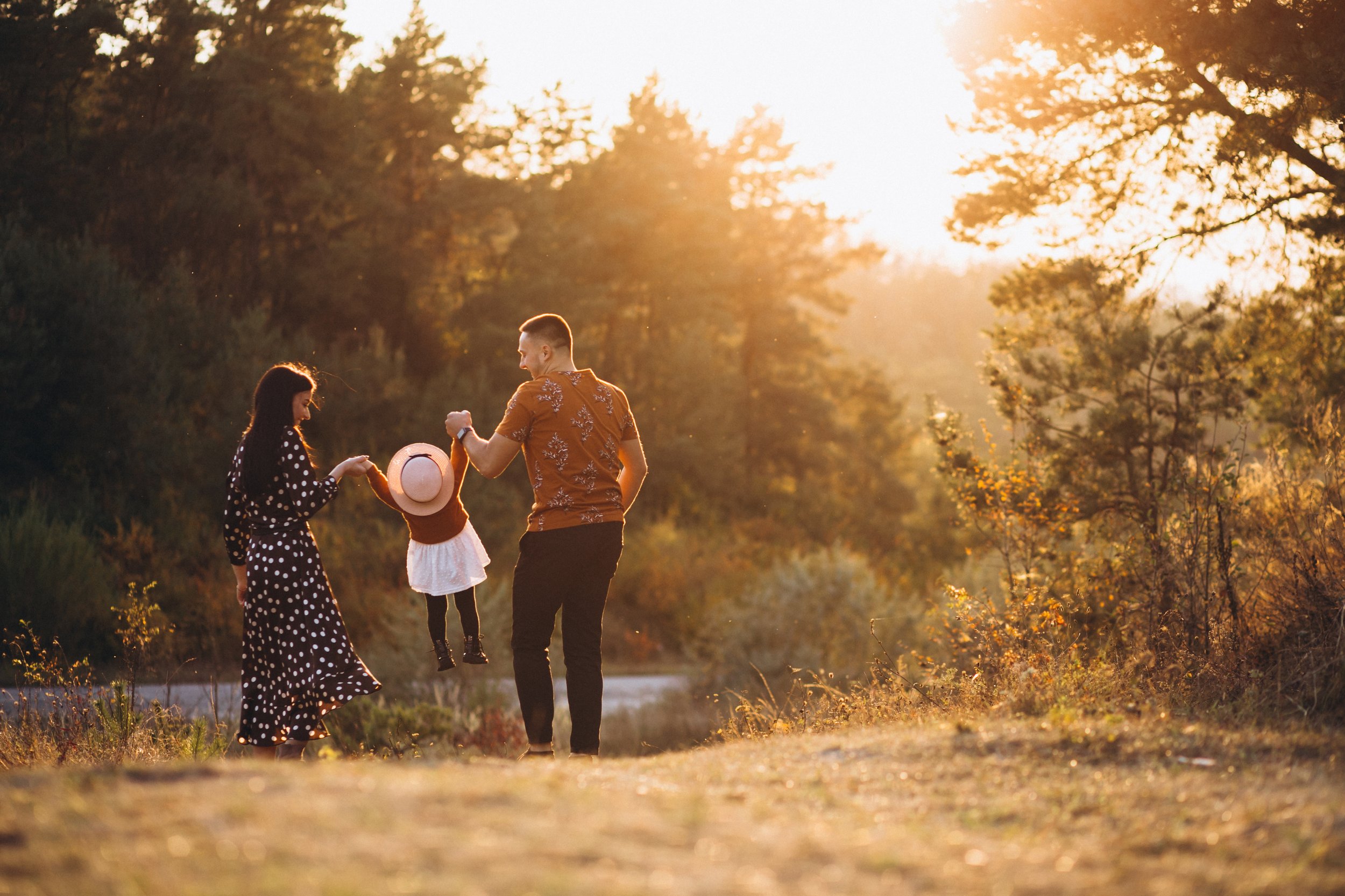 Un groupe de trois personnes, deux adultes et un enfant, jouant avec une poupée en plein air au coucher du soleil dans un environnement naturel avec des arbres et un ciel doré.
