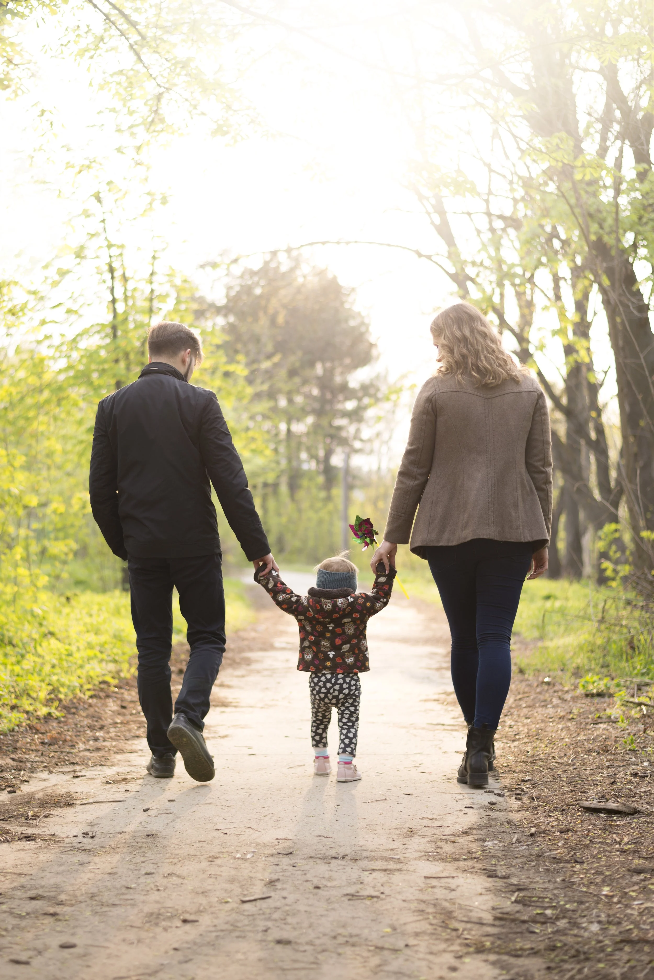Une famille avec deux adultes et un jeune enfant marche dans un chemin forestier au coucher du soleil, la mère tenant des fleurs.