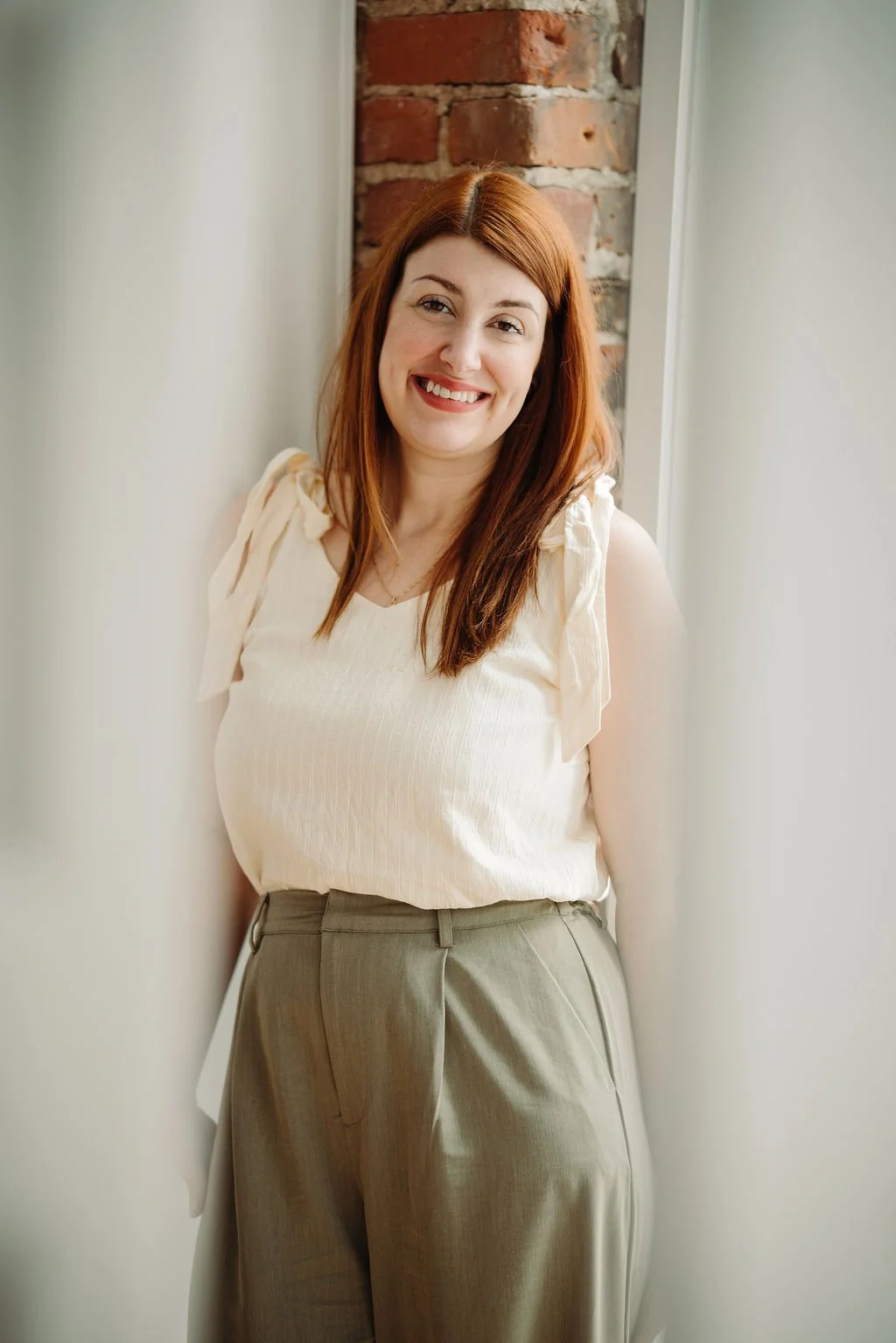 Femme souriante avec cheveux roux, portant un haut beige et pantalon beige, debout près d'un mur en pierre et d'une fenêtre, intérieur lumineux.