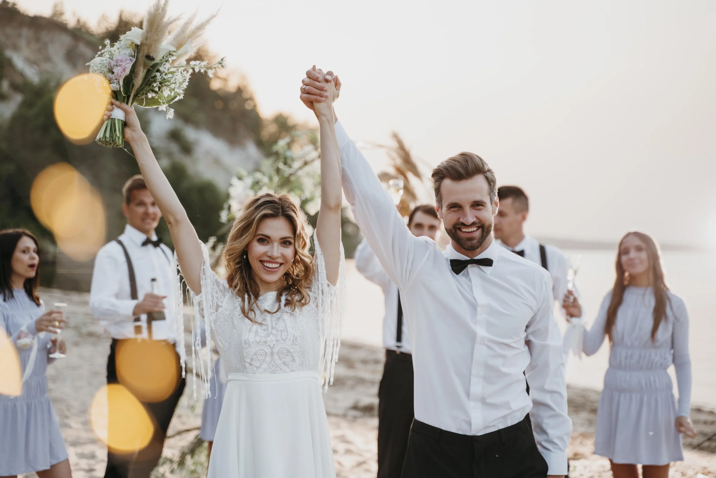 Un couple de mariés heureux à la plage entouré d'amis, célébrant leur mariage en levant les mains, avec un décor de coucher de soleil et des fleurs.