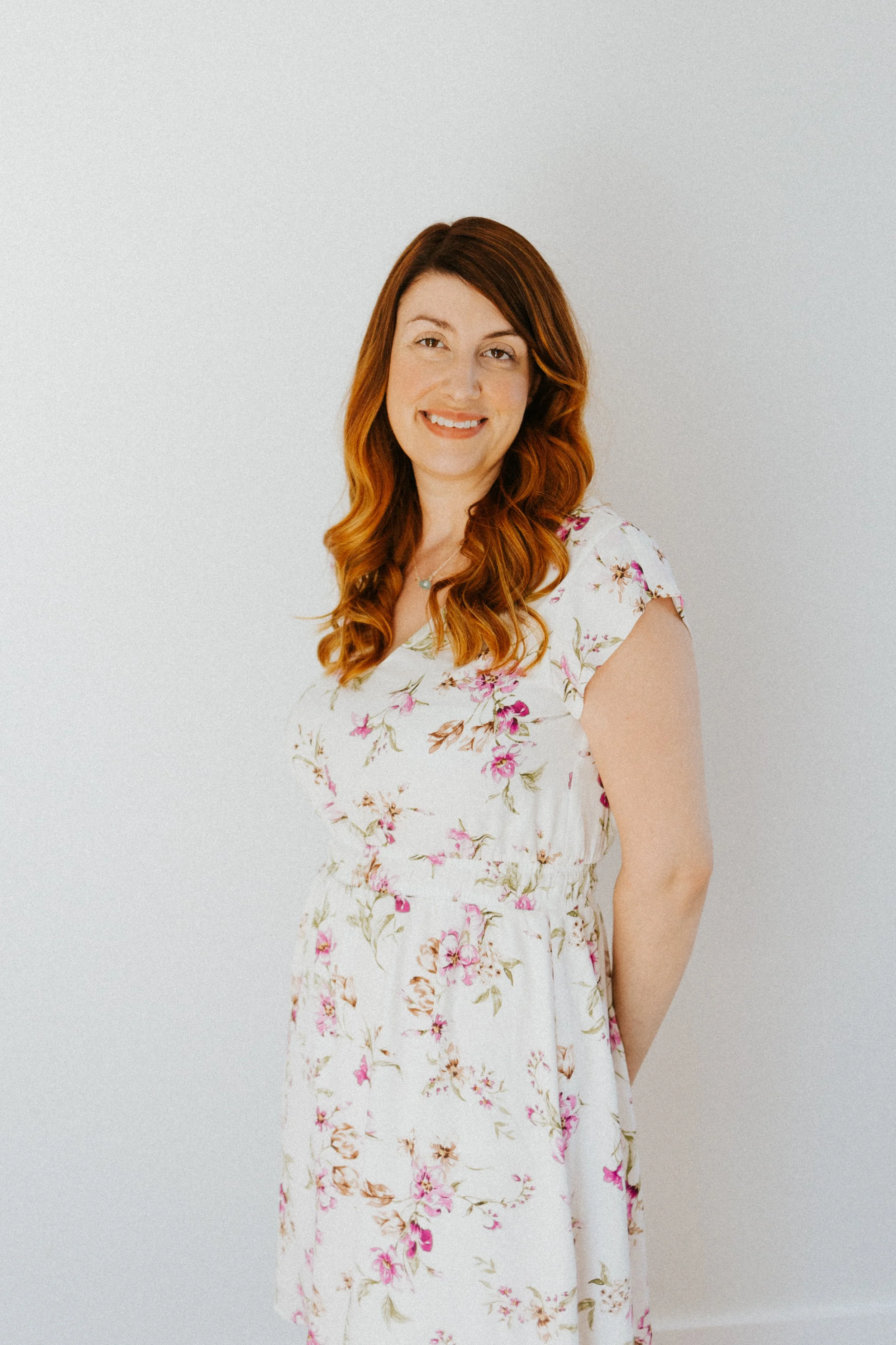 Une femme aux cheveux roux, portant une robe blanche à motifs floraux roses, souriant, debout contre un mur blanc.