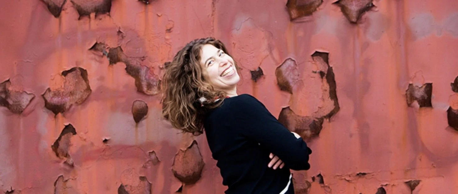 Smiling woman with curly hair posing against a textured rust-colored wall.