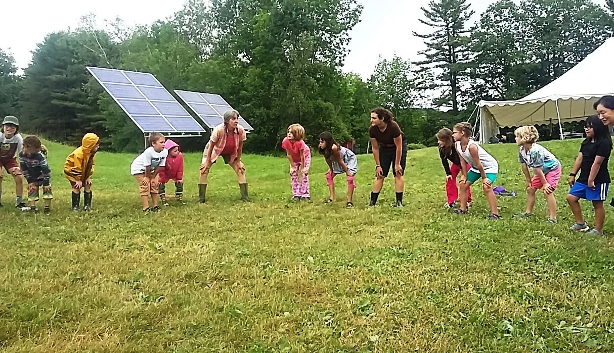 A group of children and adults bending over in a grassy field, with solar panels and a tent in the background. They appear to be participating in an outdoor activity.