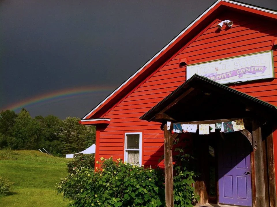 Red building with a purple door under a dark sky, featuring a rainbow in the background. A sign on the building displays a partially visible name. The foreground has lush green bushes.