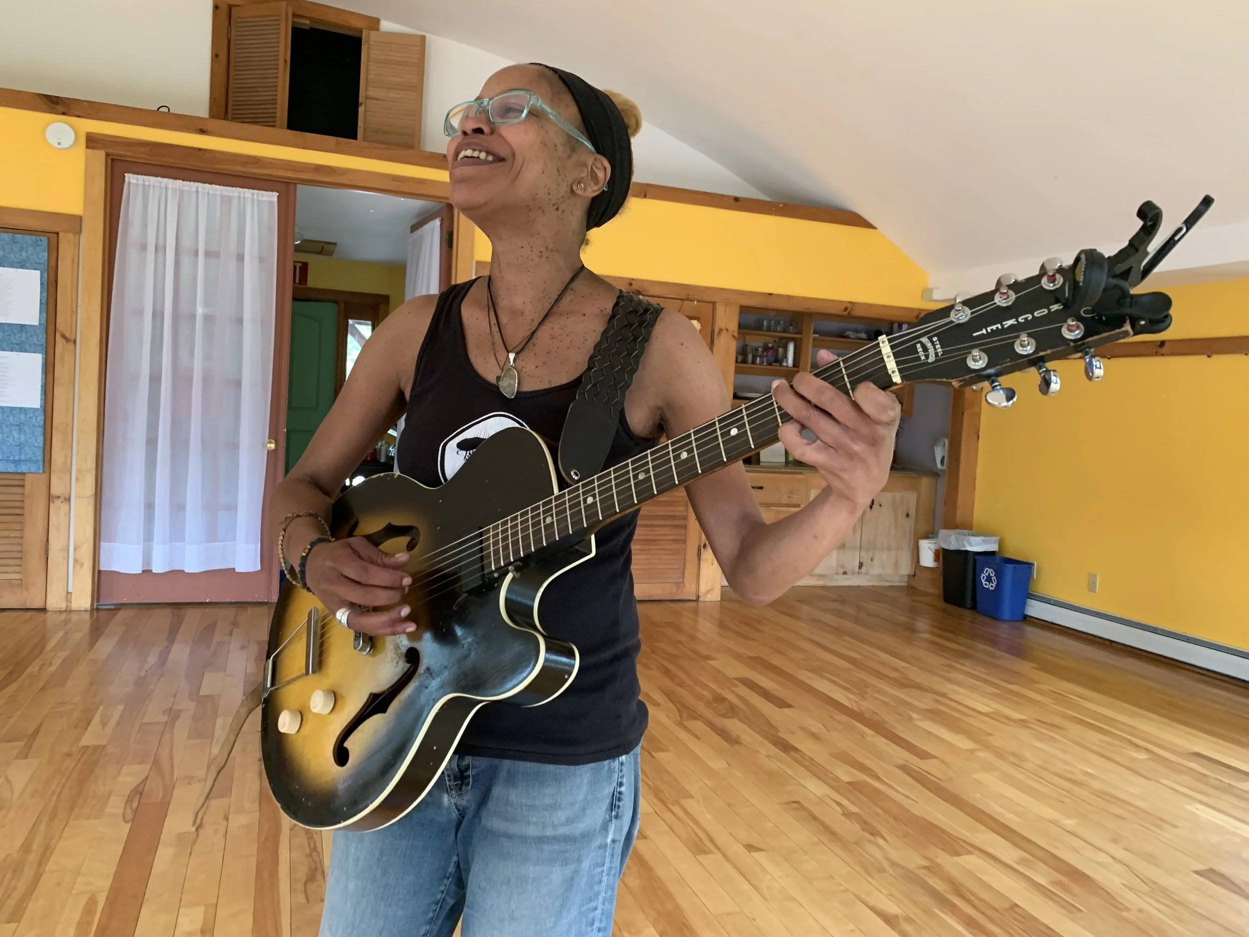 Person playing an electric guitar, smiling in a room with yellow walls and hardwood floors.