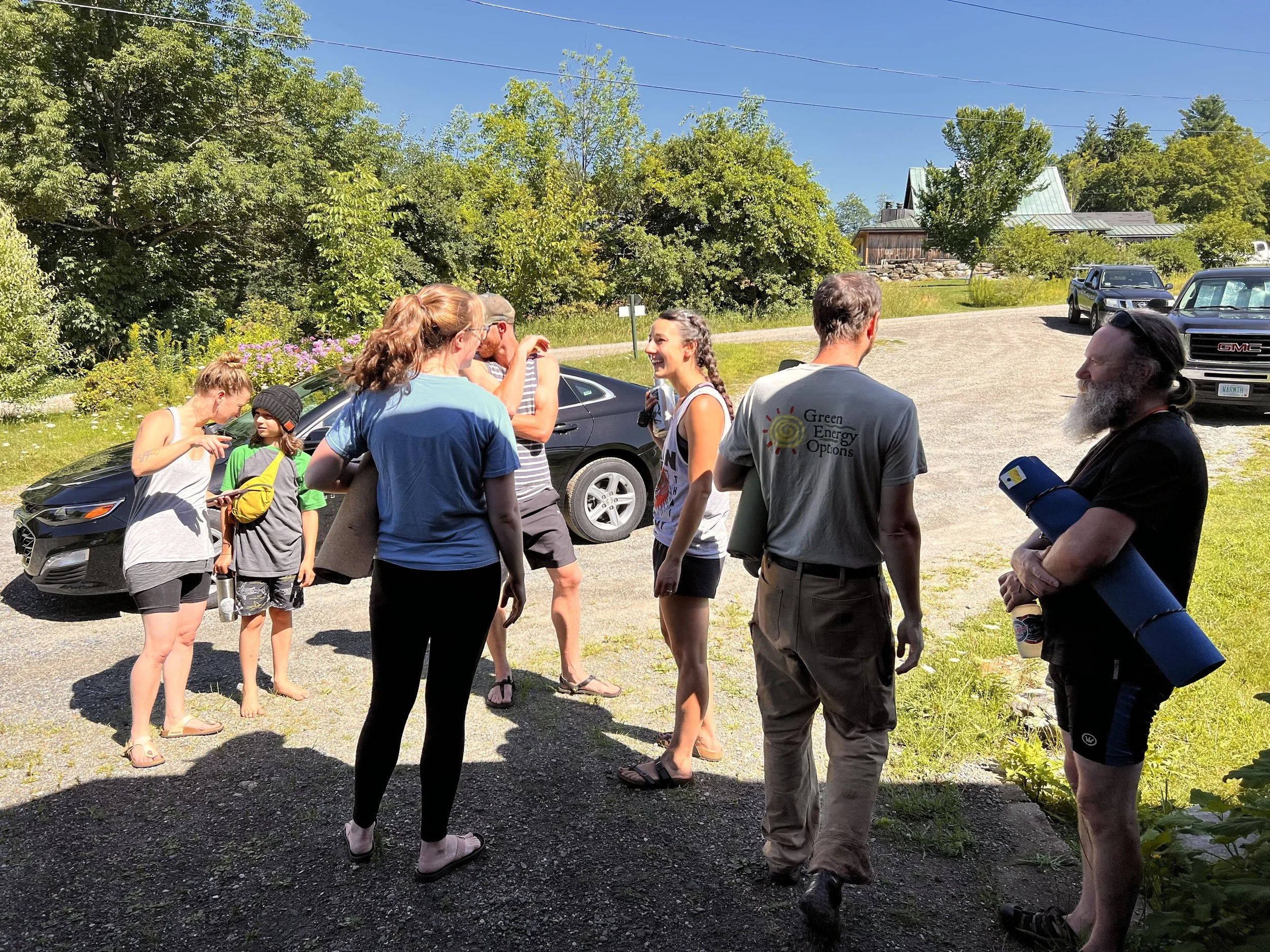 Group of people standing outside under bright blue sky, dressed casually, with one person holding a yoga mat. Cars and greenery can be seen in the background.