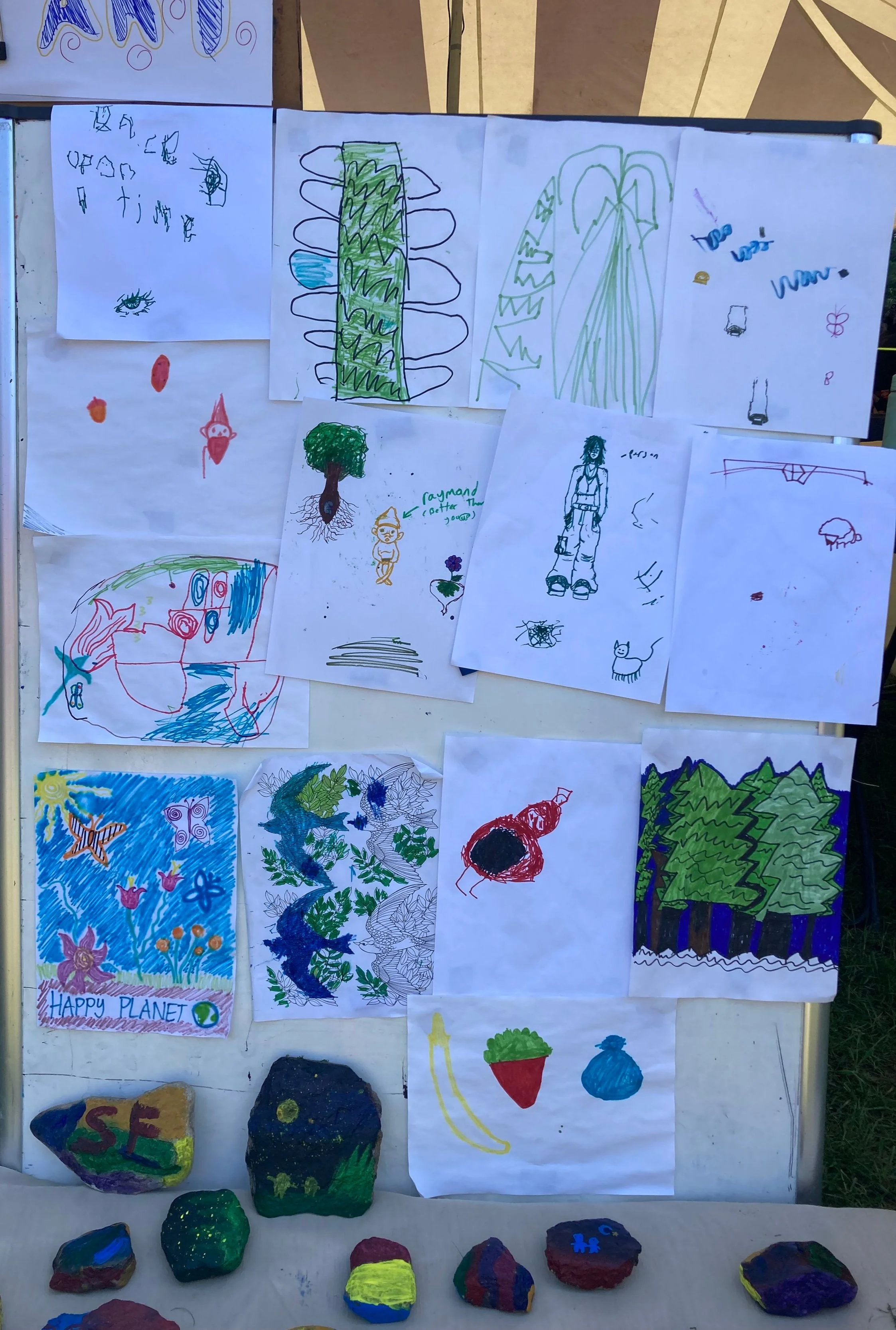 A collection of children's drawings displayed on a board, featuring colorful abstract shapes, animals, trees, and imaginative landscapes, along with hand-painted rocks in various colors placed at the bottom.
