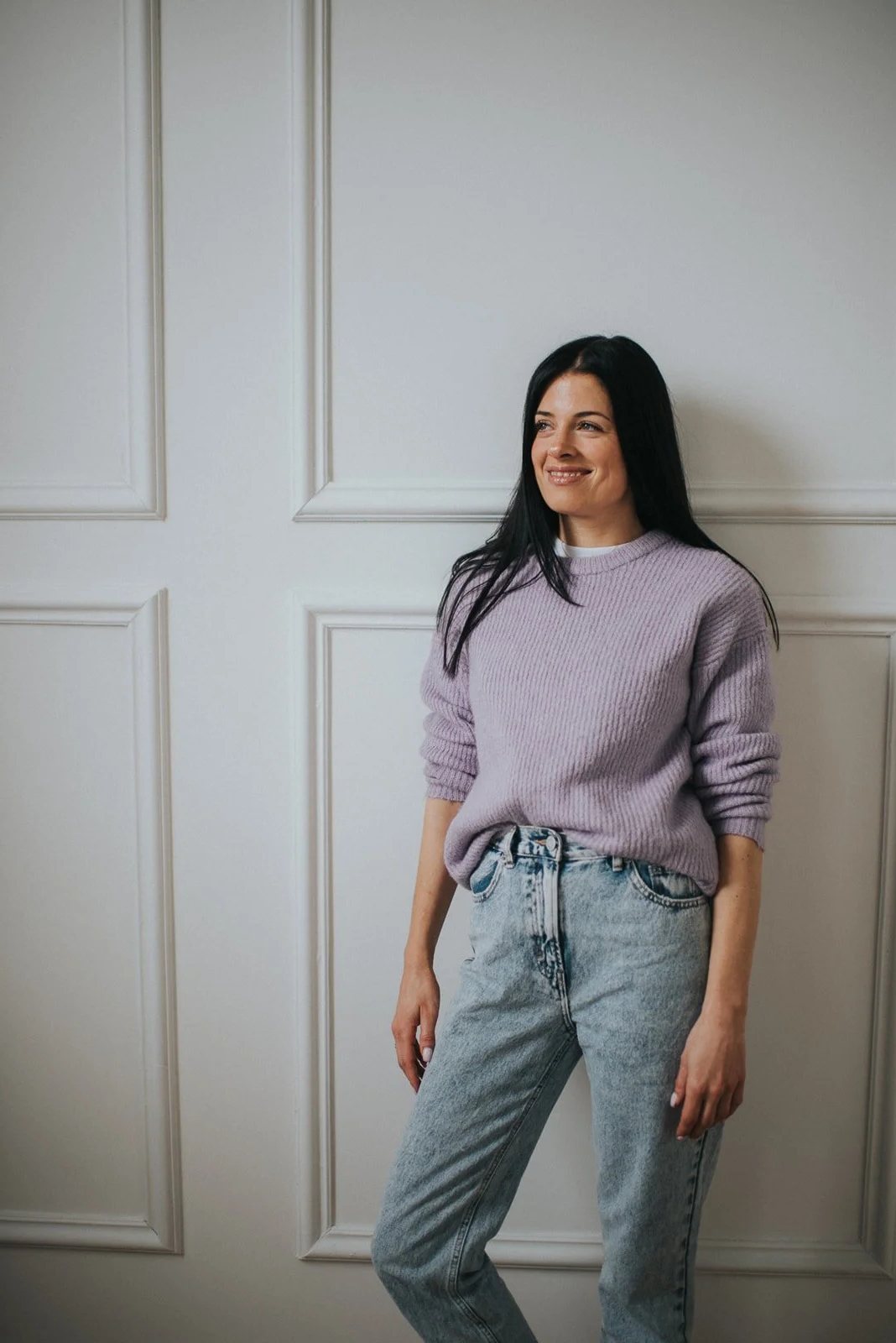 A woman with long black hair wearing a light purple sweater and light blue jeans, standing against a cream-colored paneled wall.