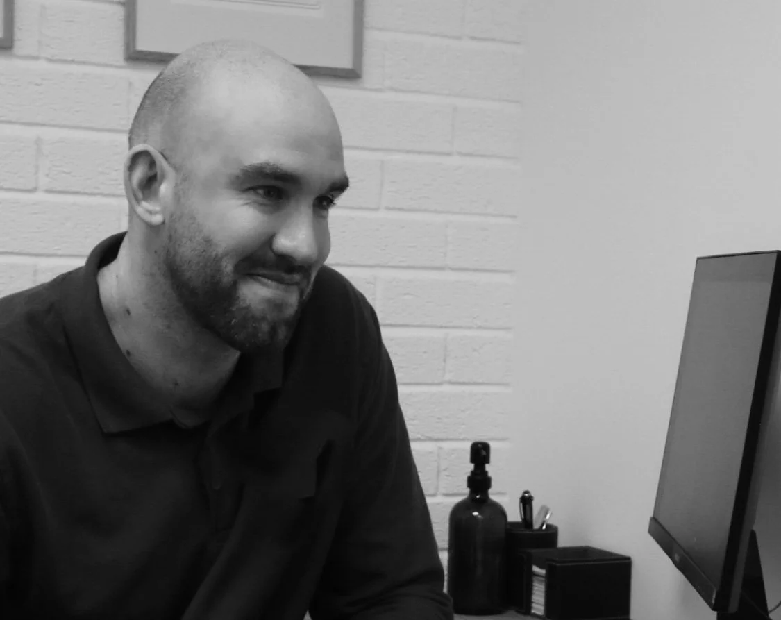 A man with a beard and a shaved head sitting at a desk, looking at a computer monitor, in an office with a brick wall behind him.