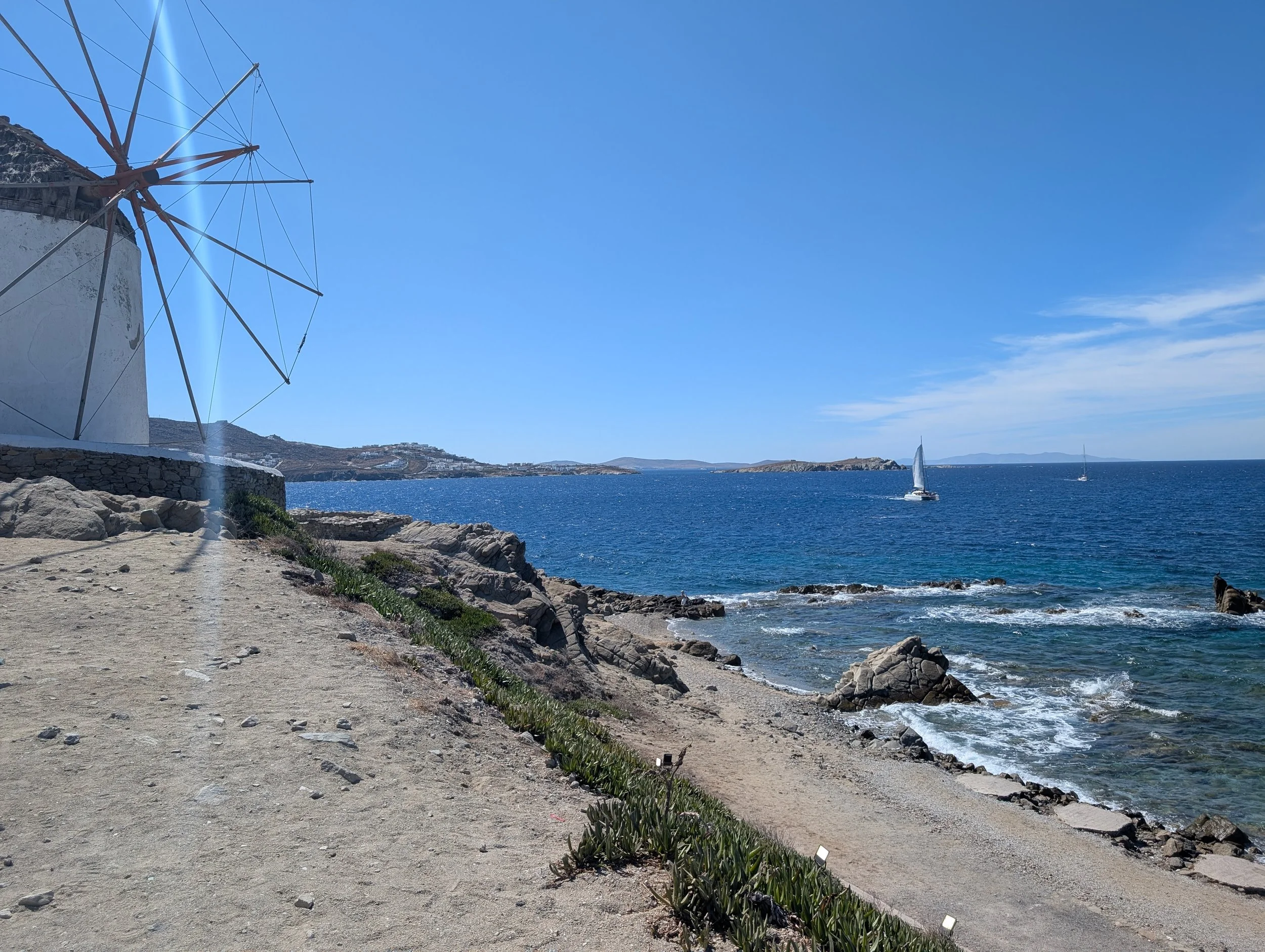 Mykonos Windmilk overlooking the beach with waves, sail boats, and rock outcroppings