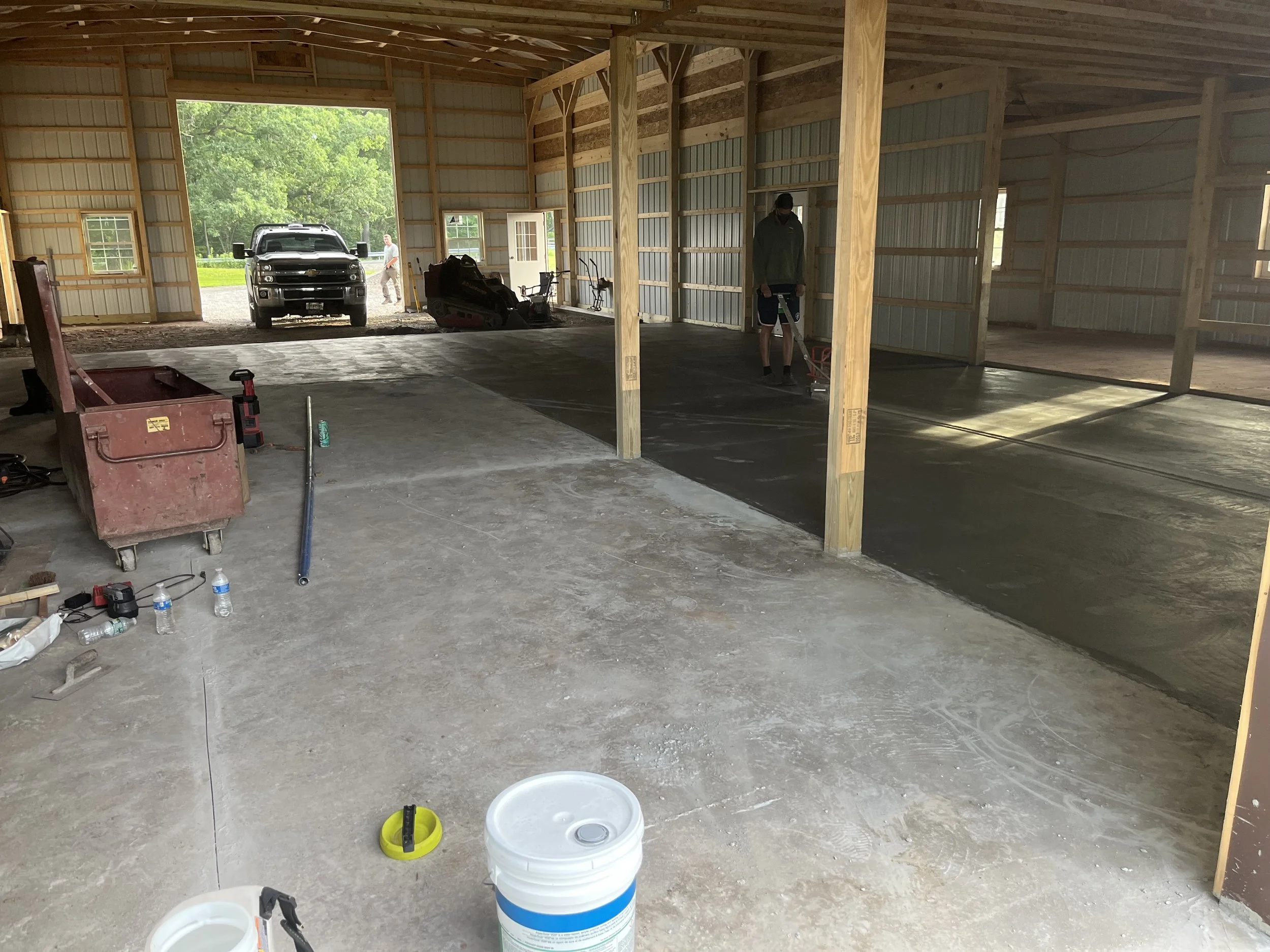 Inside a partially constructed garage with exposed wooden framing, concrete flooring, construction tools, a red toolbox, several water bottles, a bucket, and two people working on floor installation.