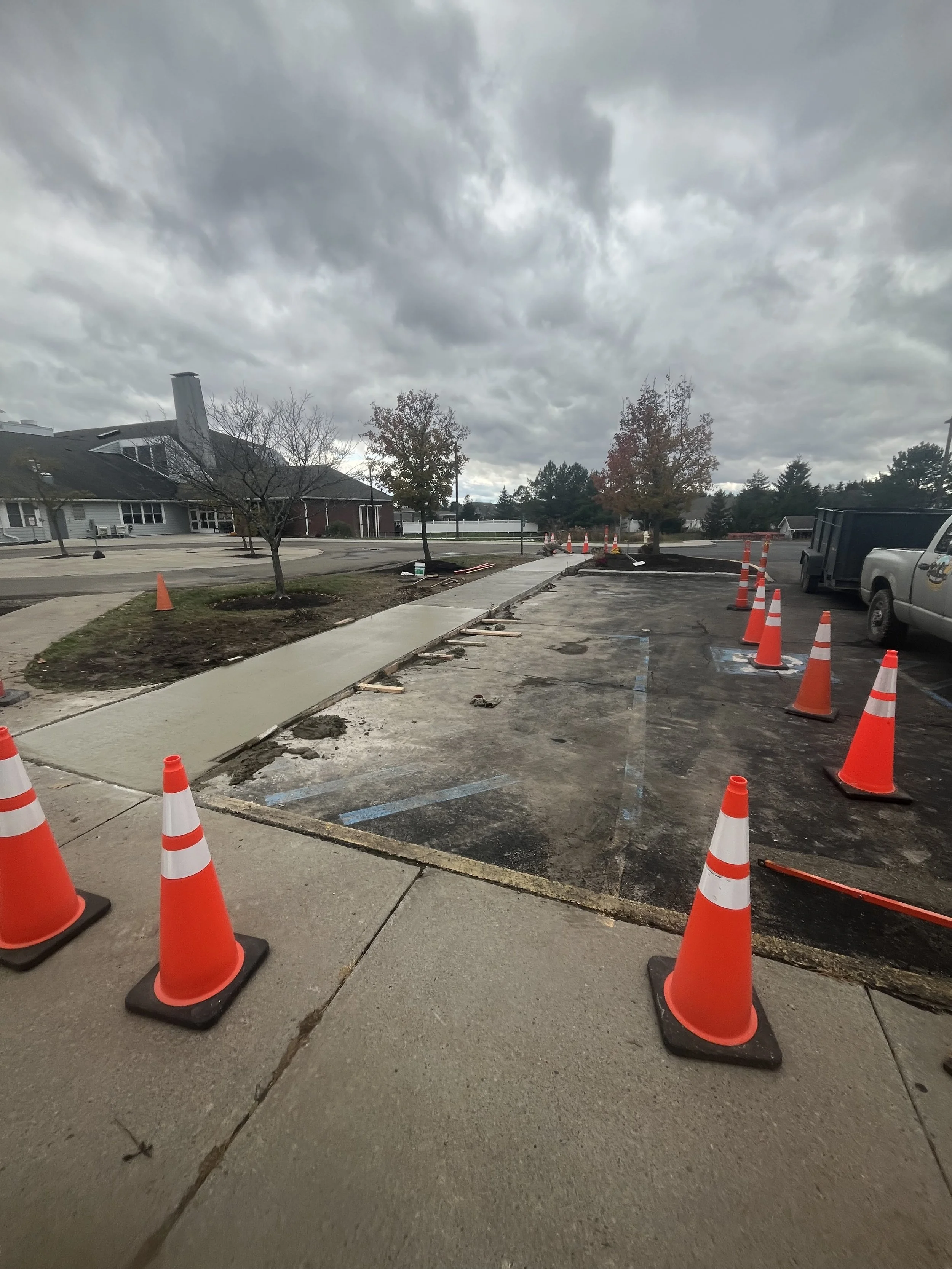 A sidewalk and parking lot with orange construction cones and ongoing pavement work on a cloudy day.
