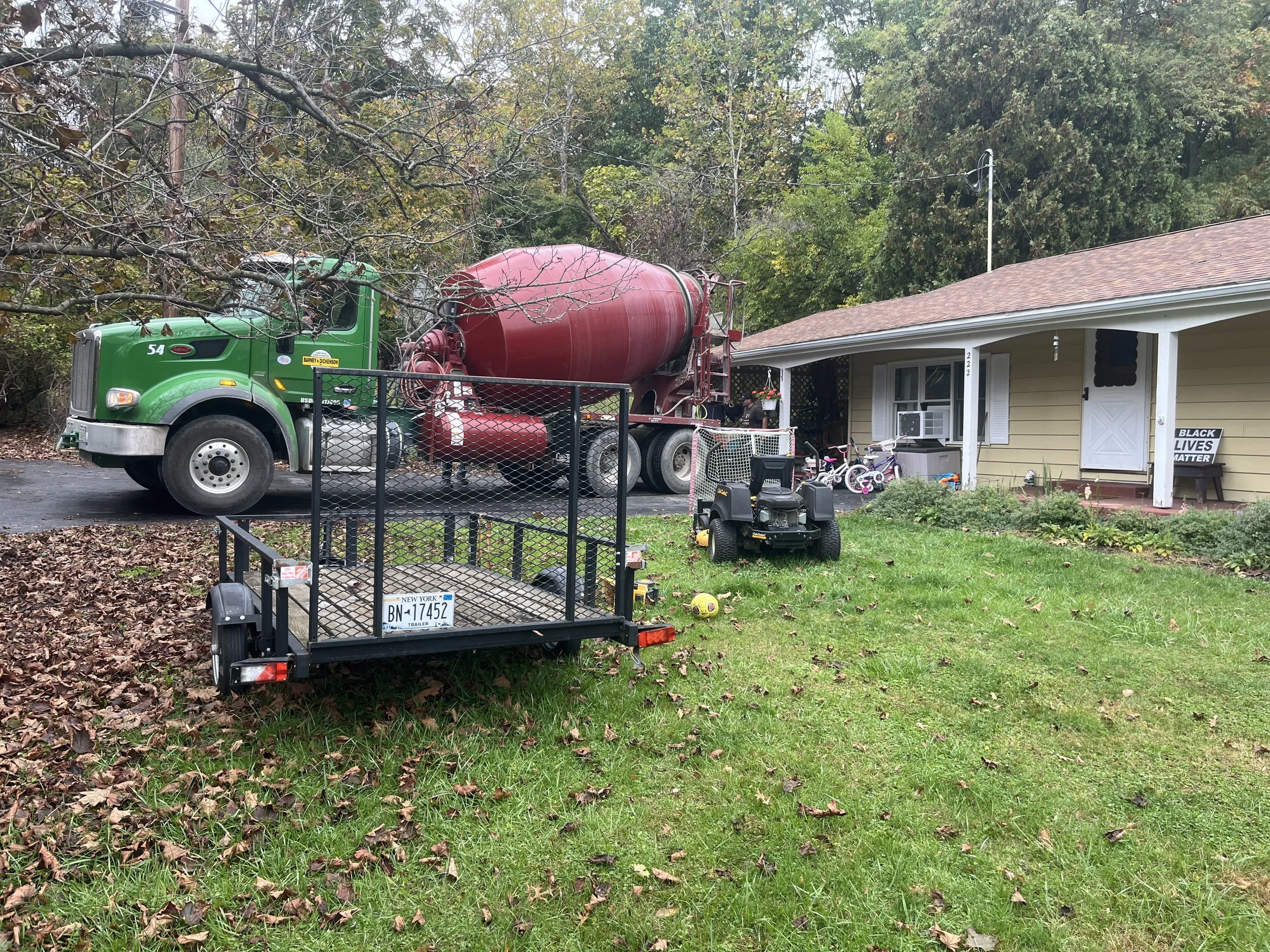 Front yard with green cement mixer truck parked on driveway, a toy trailer, children's bicycles, a small riding lawn mower, and a yellow house with a porch. There is a sign saying 'Black Lives Matter' near the porch.