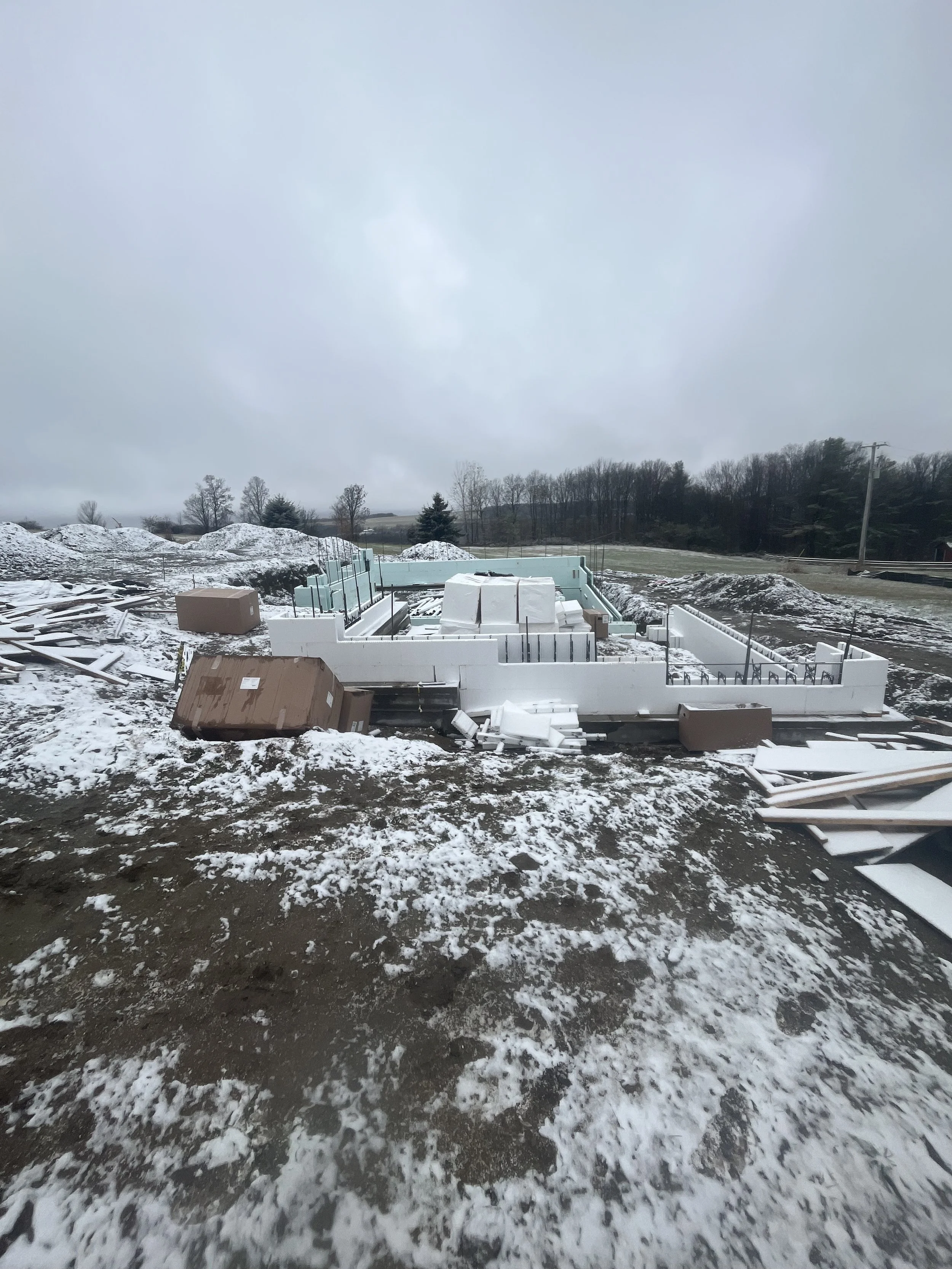 Construction site with a building foundation in progress, surrounded by snow and scattered construction materials, under an overcast sky.