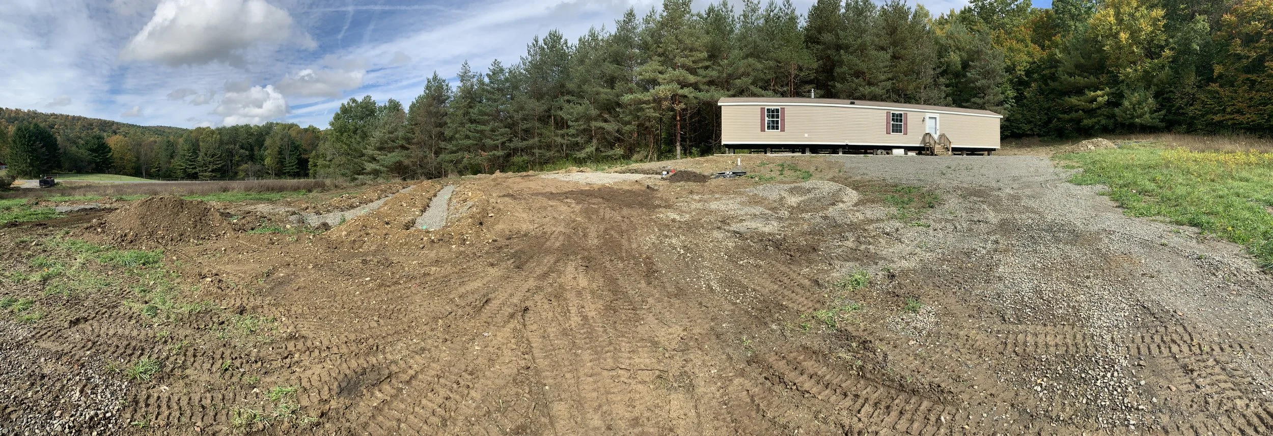 Construction site with dirt and gravel, mobile home in the background, surrounded by trees and a partly cloudy sky.