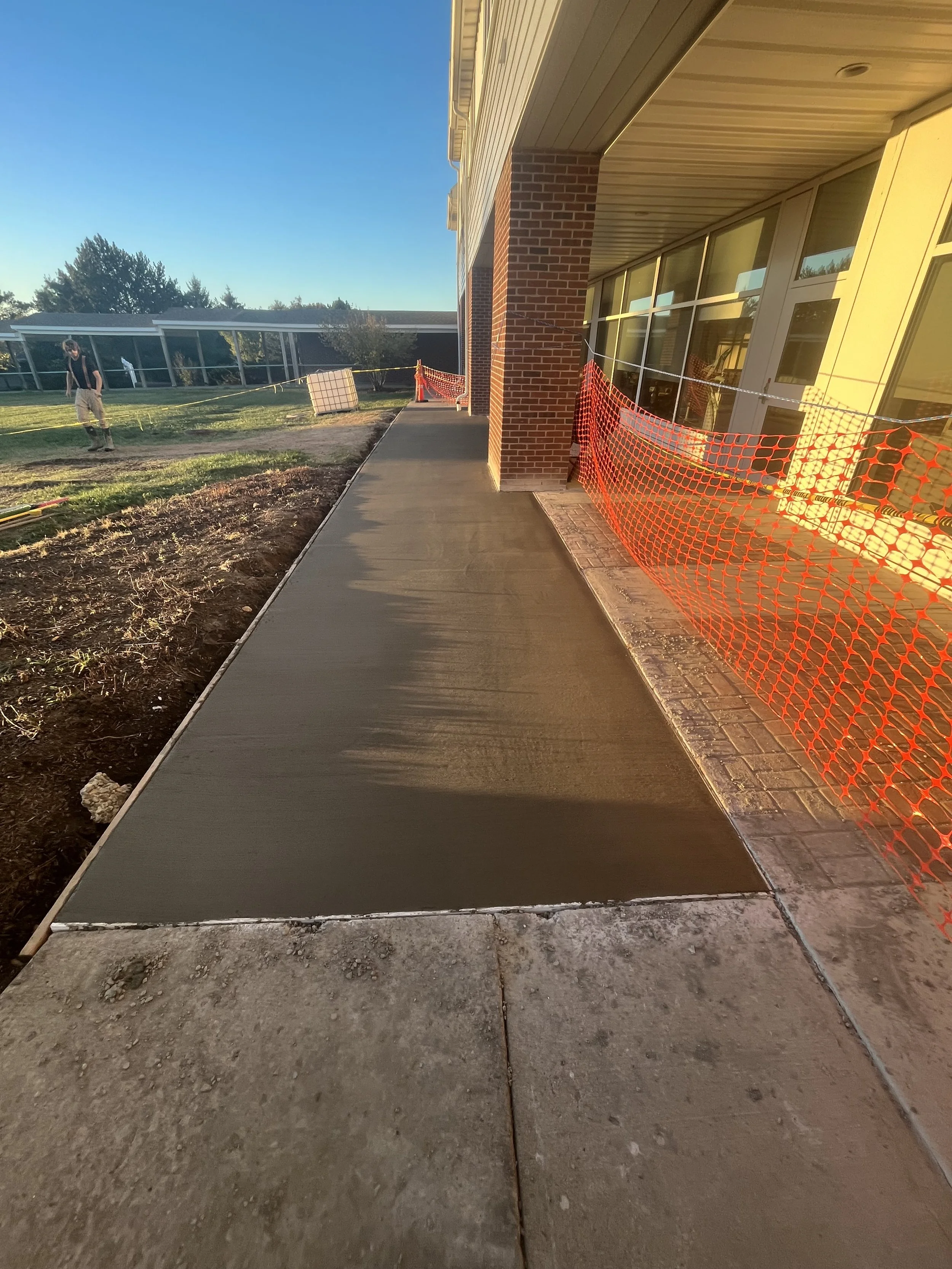 Freshly poured concrete sidewalk outside a building with red brick wall and large glass windows, orange construction safety fencing on one side, workers in the background under an open sky.