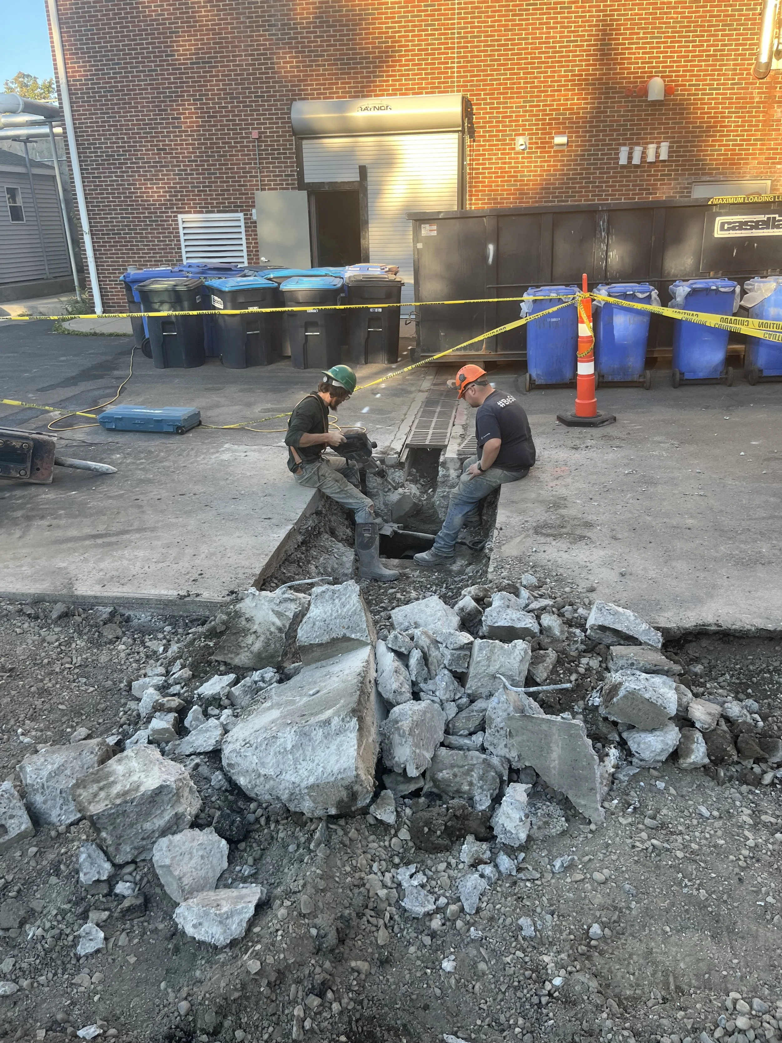 Two construction workers in safety helmets working on a street with broken pavement, surrounded by debris, with a row of trash bins and a brick building in the background.