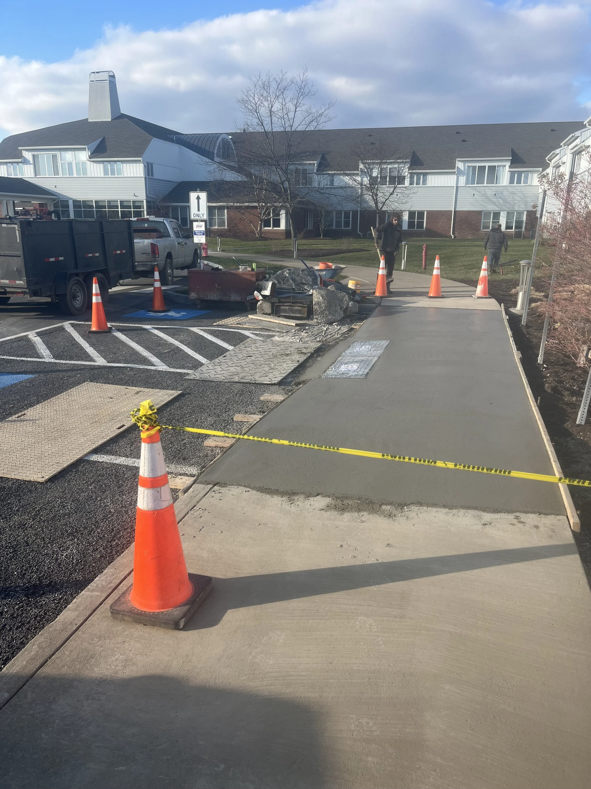 Construction workers paving a sidewalk with orange traffic cones and yellow caution tape, in front of a residential building under a partly cloudy sky.