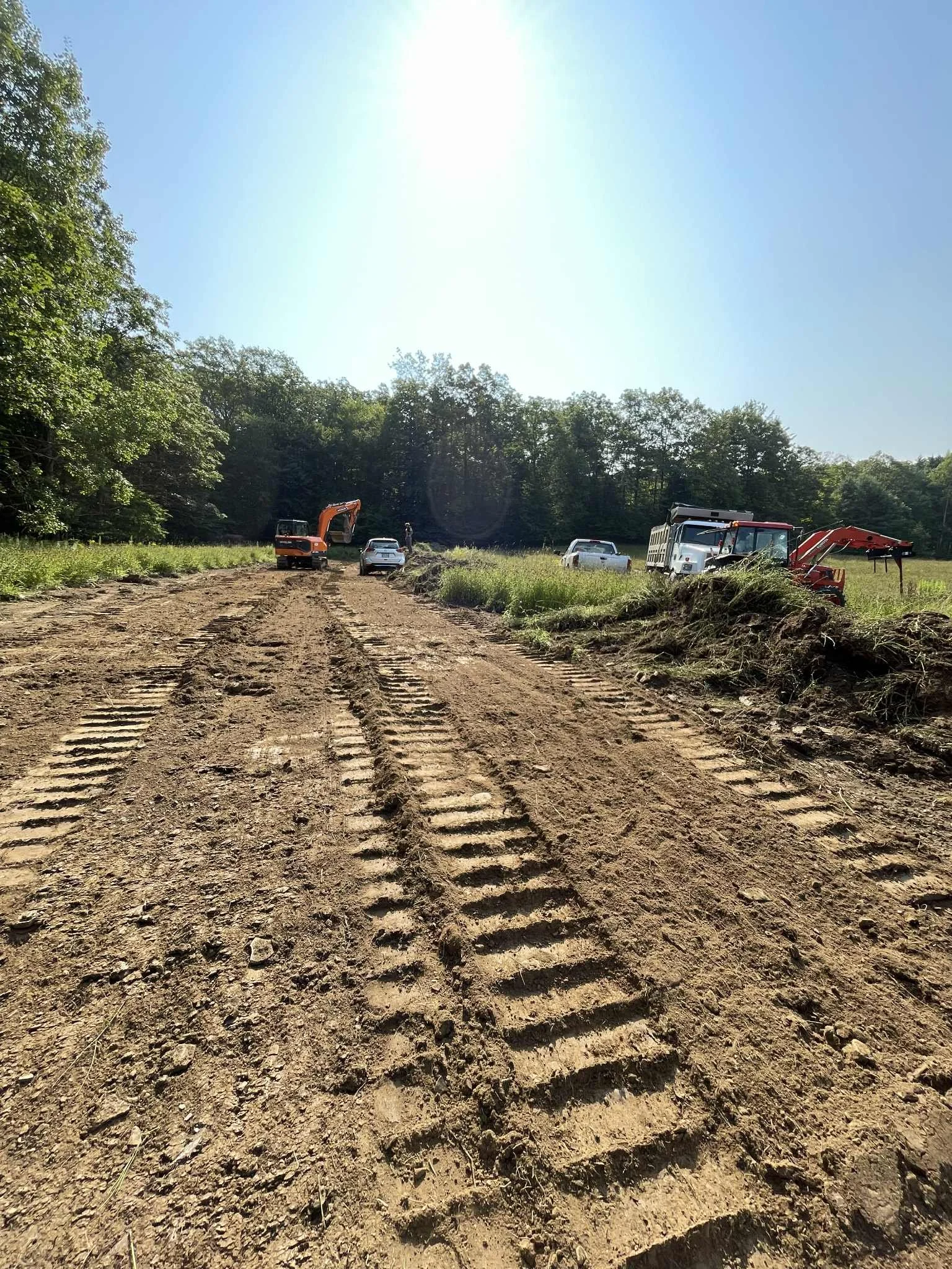 Construction site with dirt road, tracked vehicles, and construction equipment near a grassy field and trees, under a bright sunny sky.