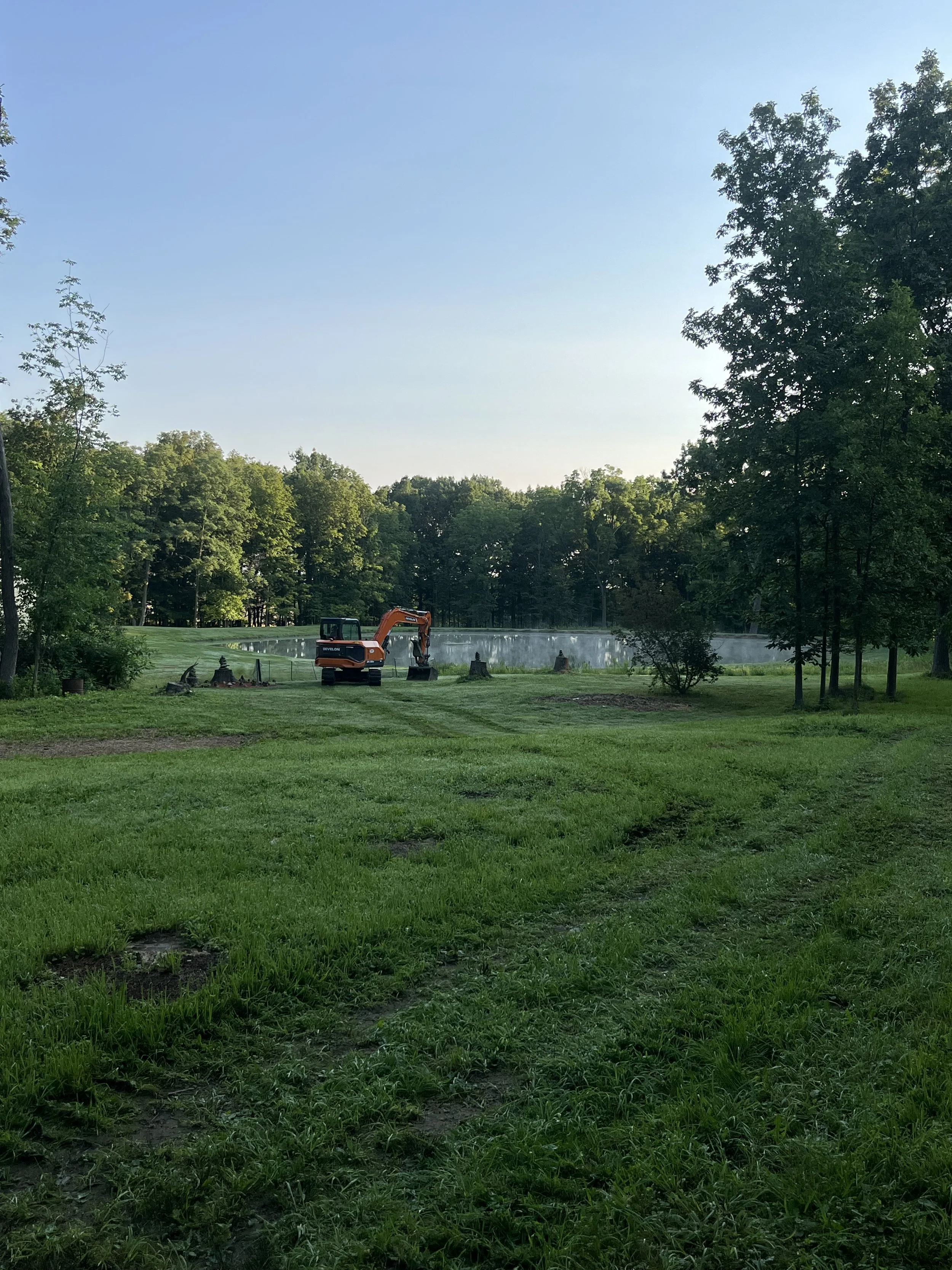 A landscape with a grassy area, trees, a pond with mist or fog, and a small orange excavator near the pond.