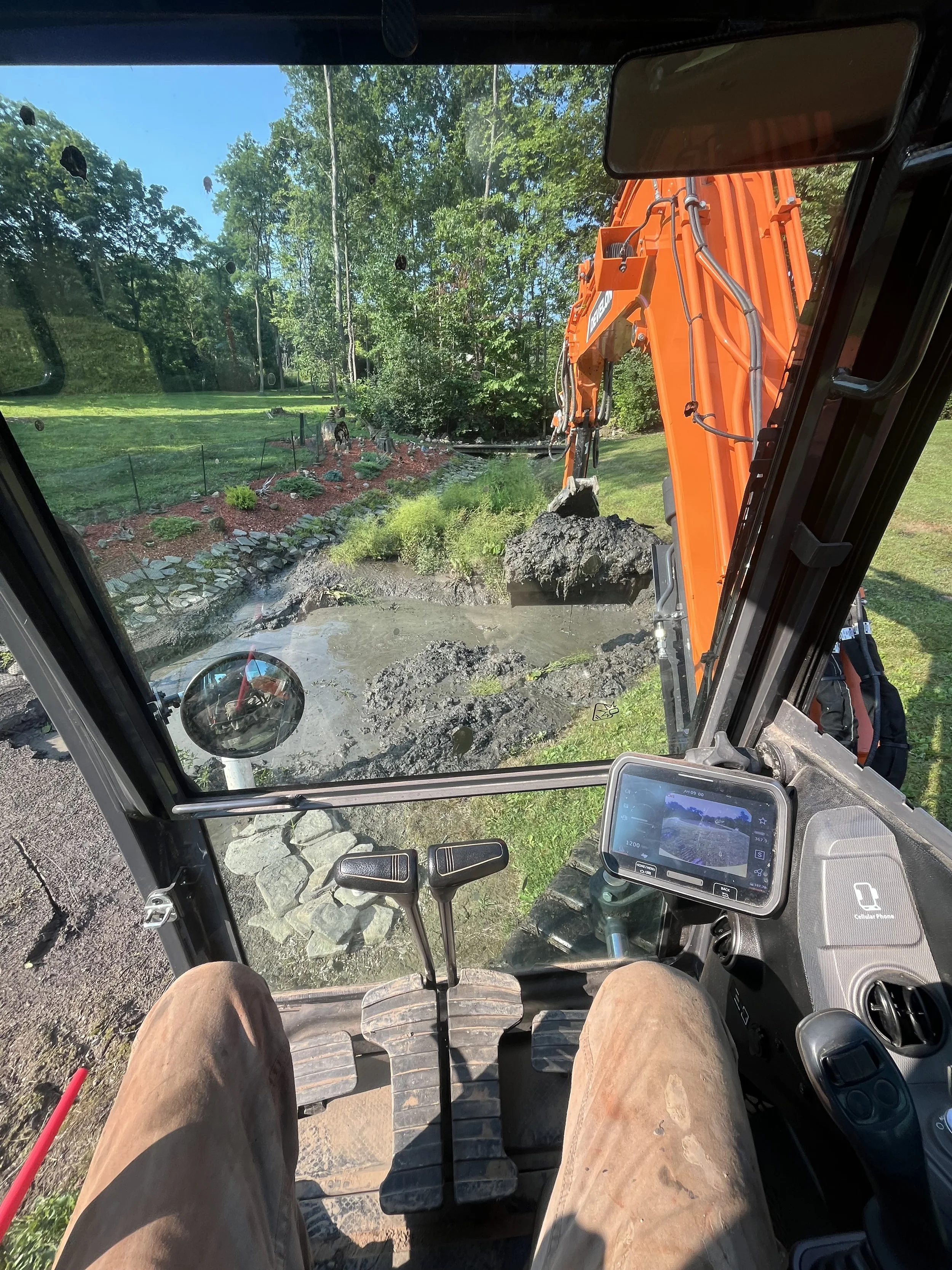 View from inside a construction excavator cabin showing a dirt digging operation with a bright orange arm digging up soil, a remote-controlled device or phone mounted inside, and a landscaped garden with trees and a lawn outside.