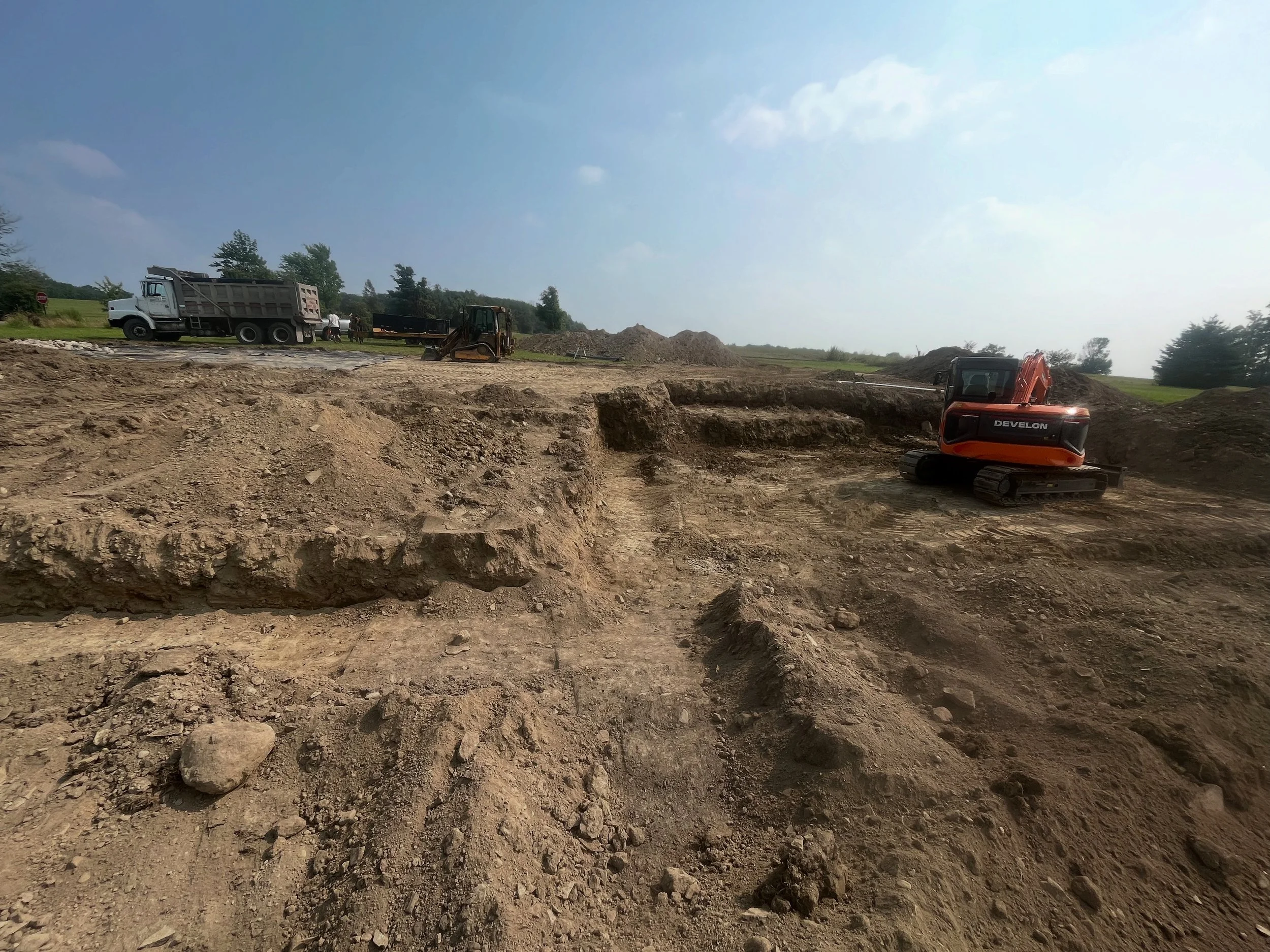 Construction site with a small orange excavator, a dump truck, and other construction equipment on a dirt lot under a partly cloudy sky.