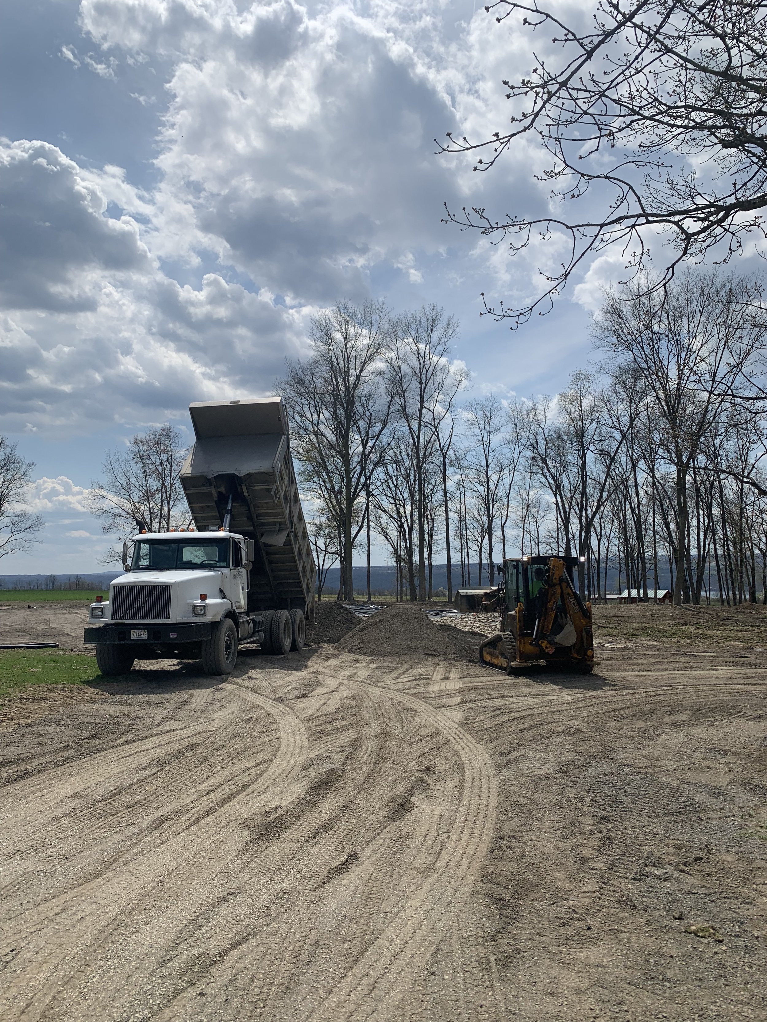 A construction site with a dump truck unloading dirt and a small excavator on a dirt road, surrounded by leafless trees under a partly cloudy sky.