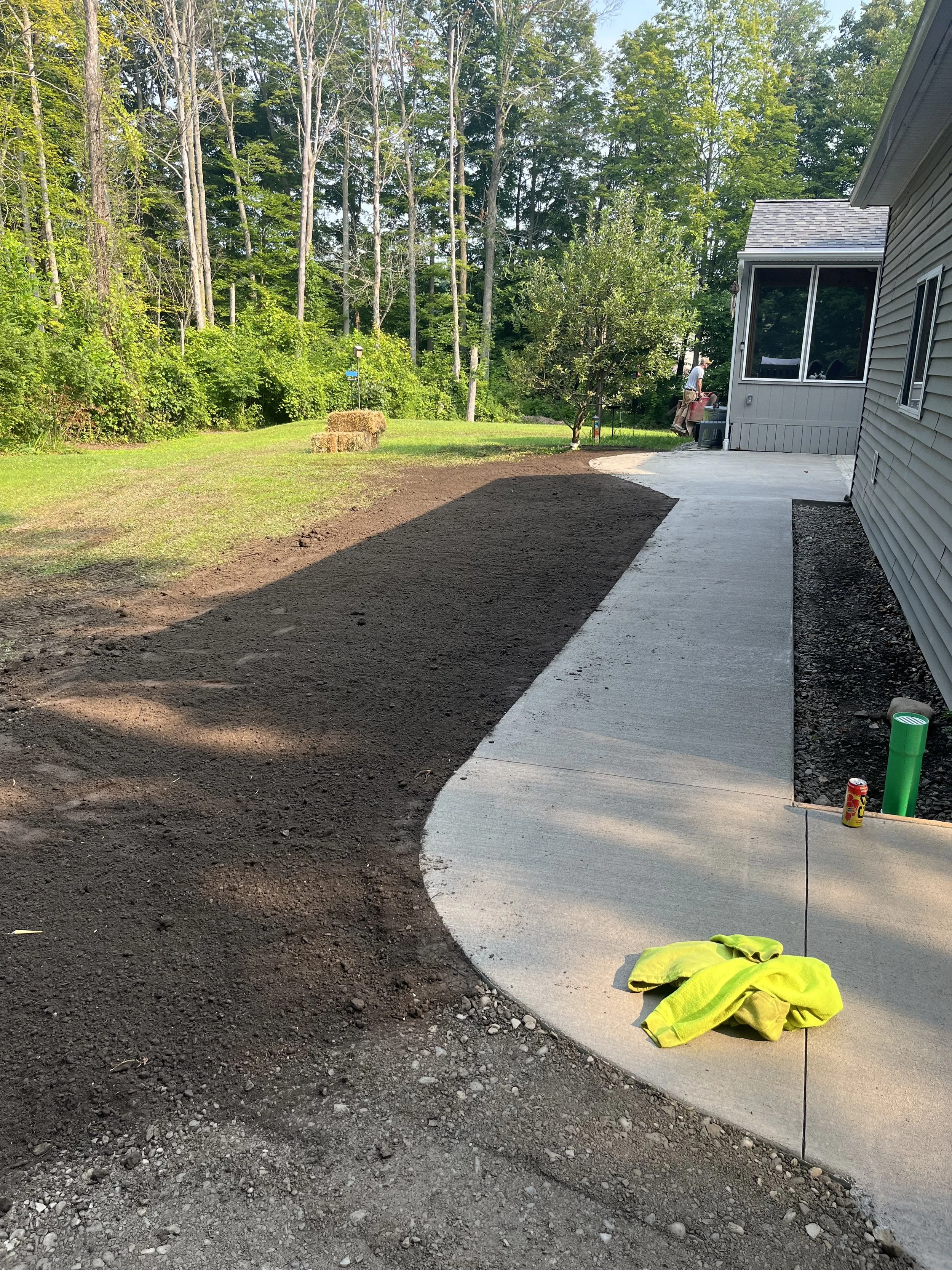A newly poured concrete sidewalk next to a house, with a section of dark soil to be landscaped. A person's reflection can be seen in the house's window, and a person in the background is standing near a tree, holding tools. Safety vest and a soda can
