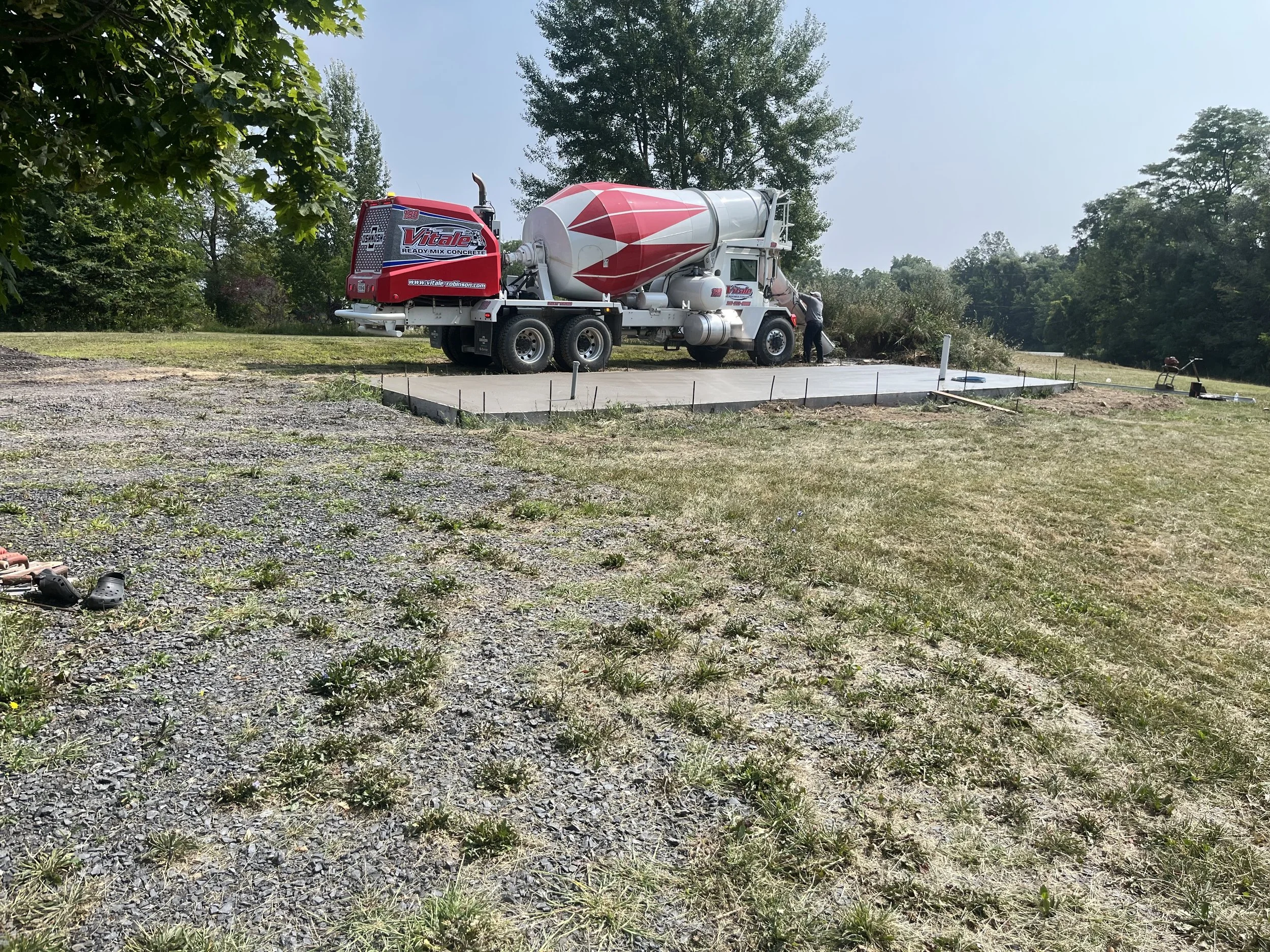 A concrete slab being poured by a mixer truck in a grassy outdoor area with trees in the background and a person operating the truck.