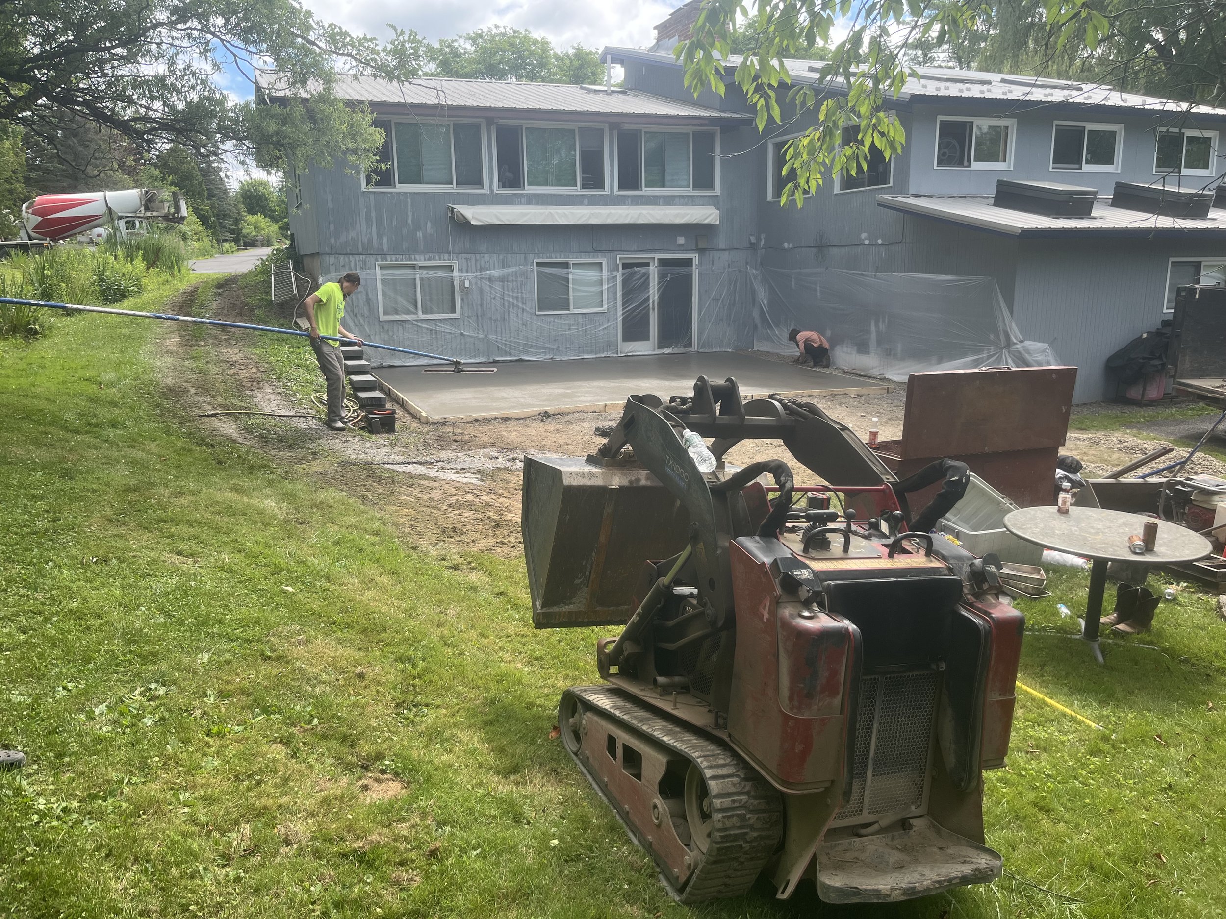 Construction workers working on a concrete patio in the backyard of a house, with construction equipment and tools visible on the grass.
