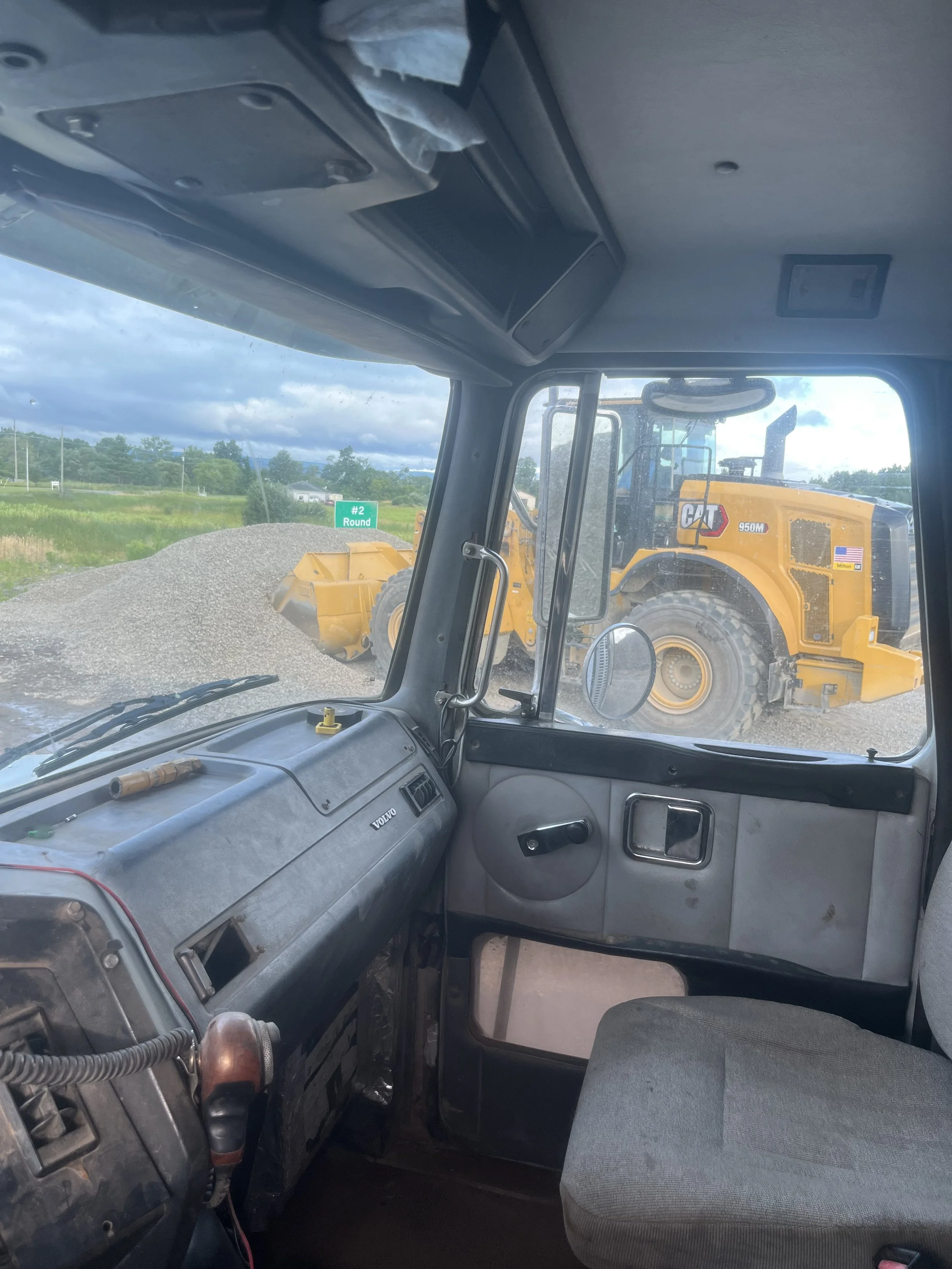 Inside the cab of a truck, view of a large Caterpillar front loader and a gravel pile outside through the windshield on a cloudy day.