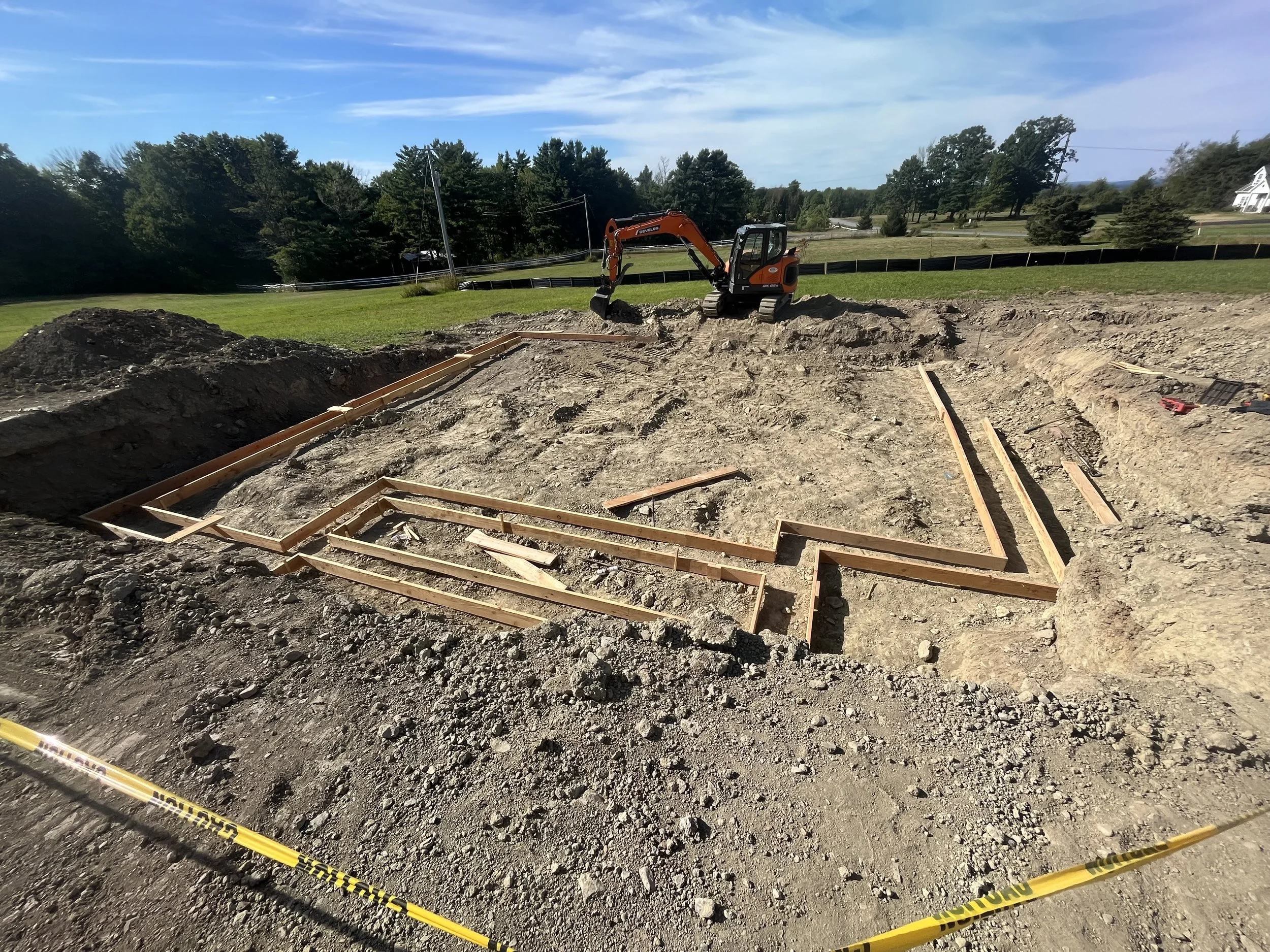 Construction site with wooden frames outlining a foundation, a small excavator, and yellow caution tape, with trees and a blue sky in the background.