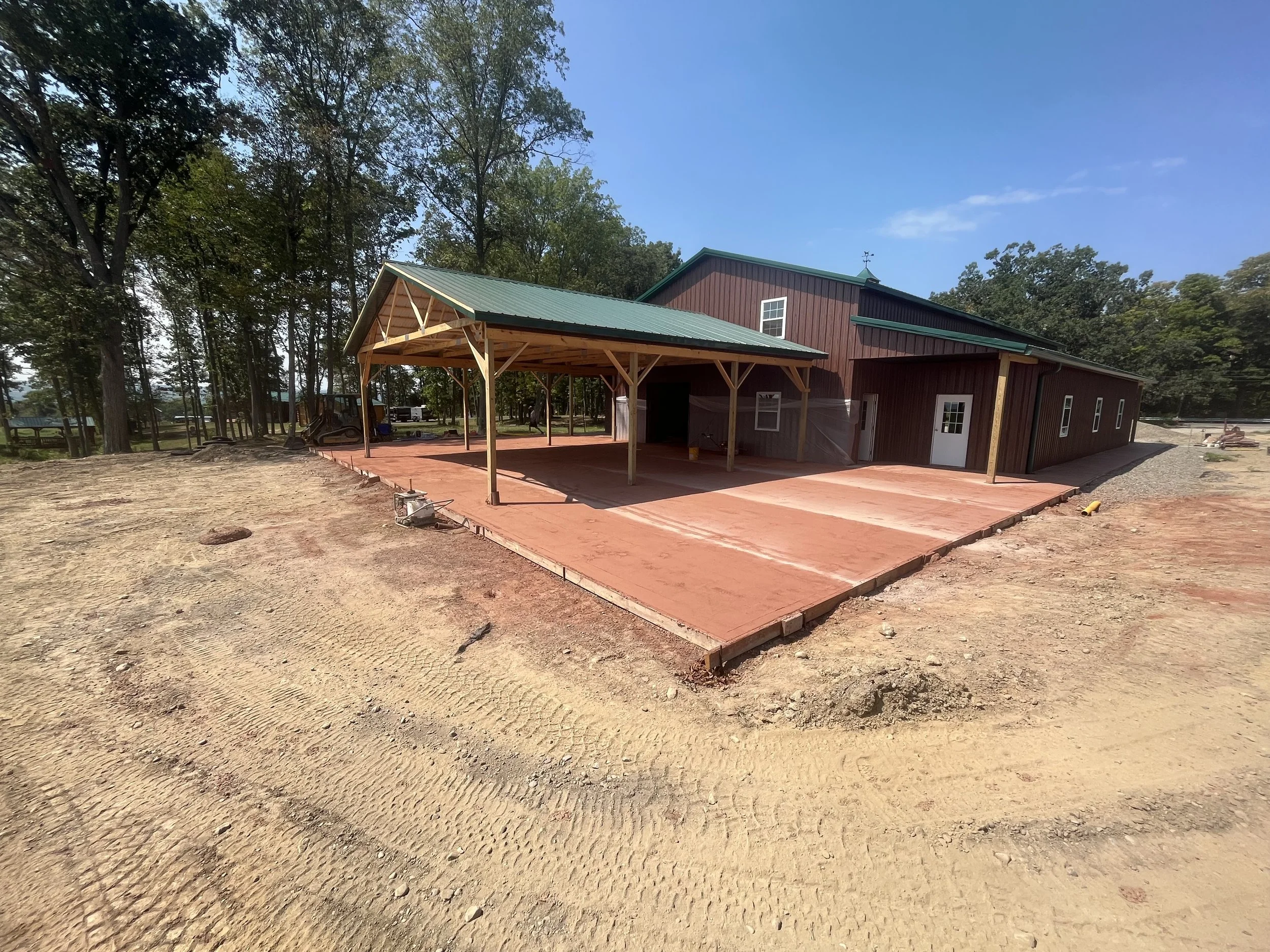 Construction site with a new building featuring a green metal roof, red wooden walls, and a large covered patio area on a sunny day.