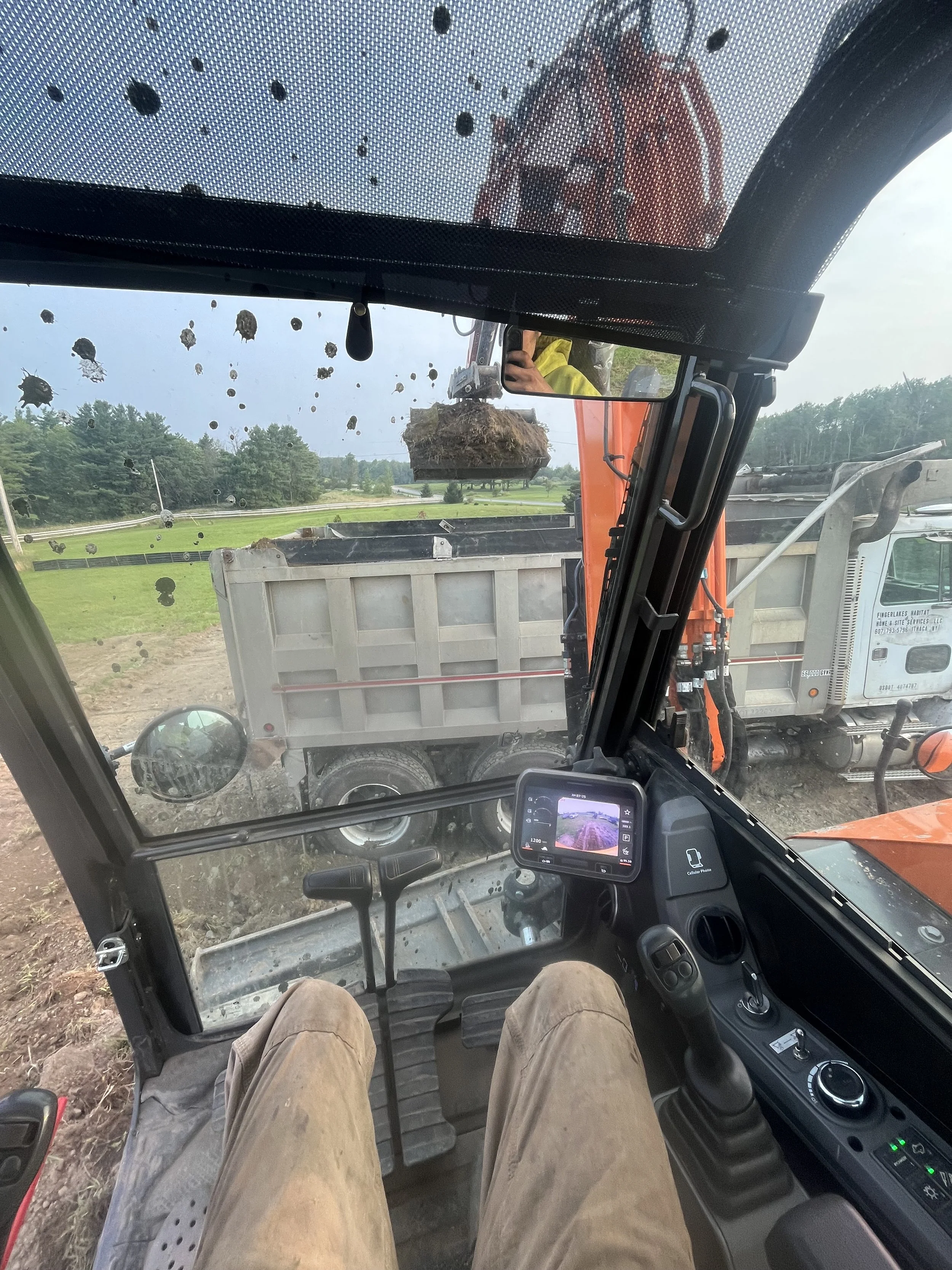 Inside the cabin of a construction vehicle, showing a person with brown pants operating the machinery, with a view of a dump truck dumping soil or mulch in front of them. The scene appears to be at a construction or landscaping site, with green field