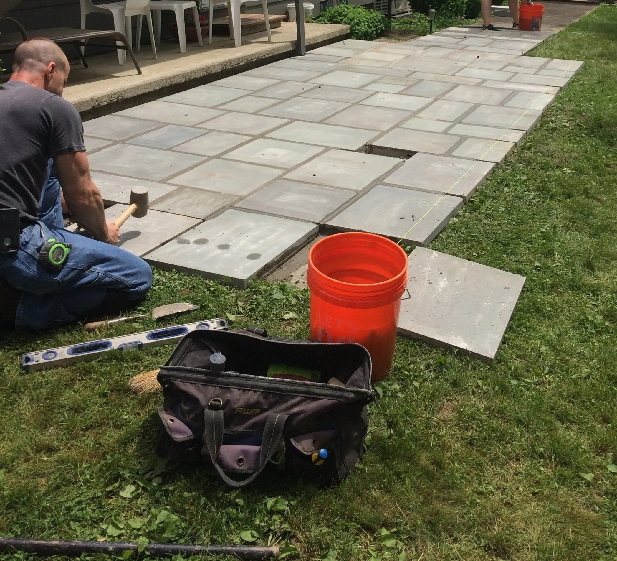 A person using a hammer to lay concrete pavers on a grassy area with a level, a backpack, an orange bucket, and a yellow string line nearby. Several pavers are already in place, with one gap visible.