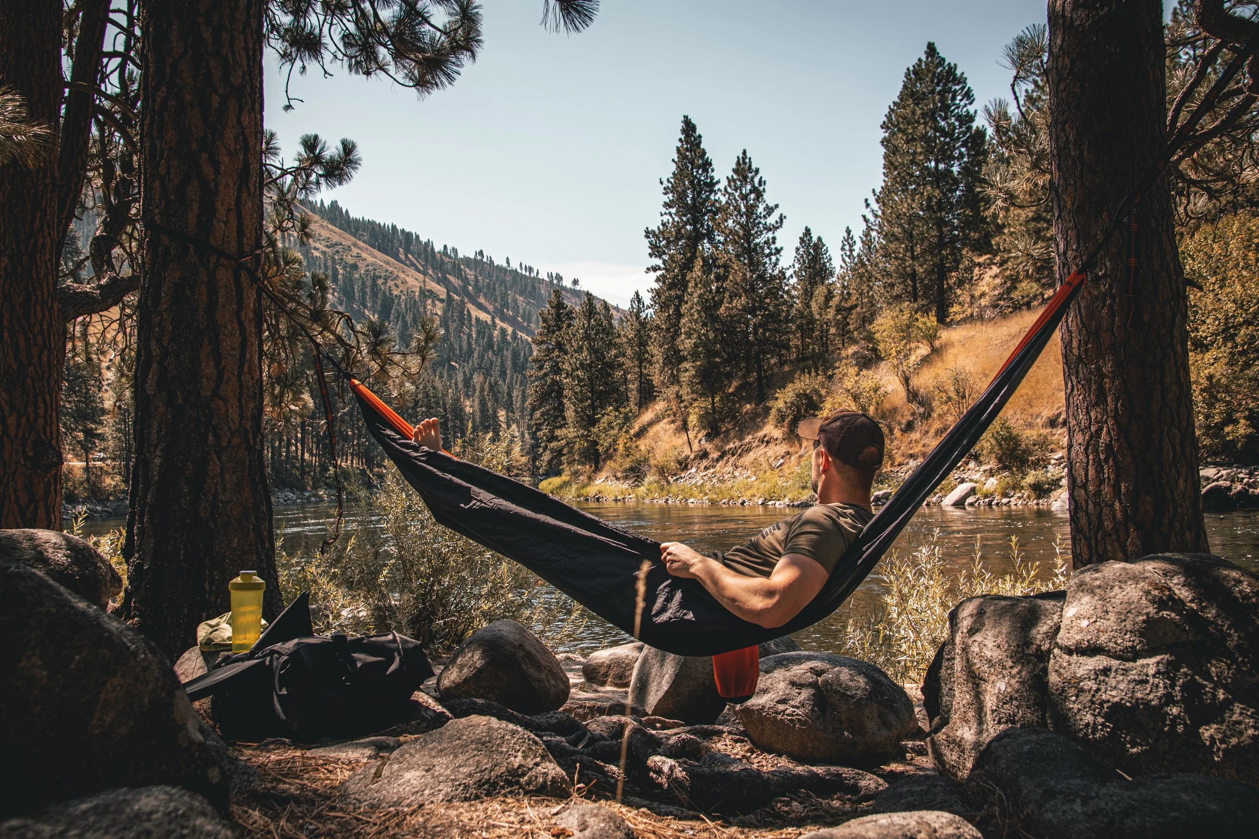 Person relaxing in a hammock by a river with trees, rocks, and mountains in the background.