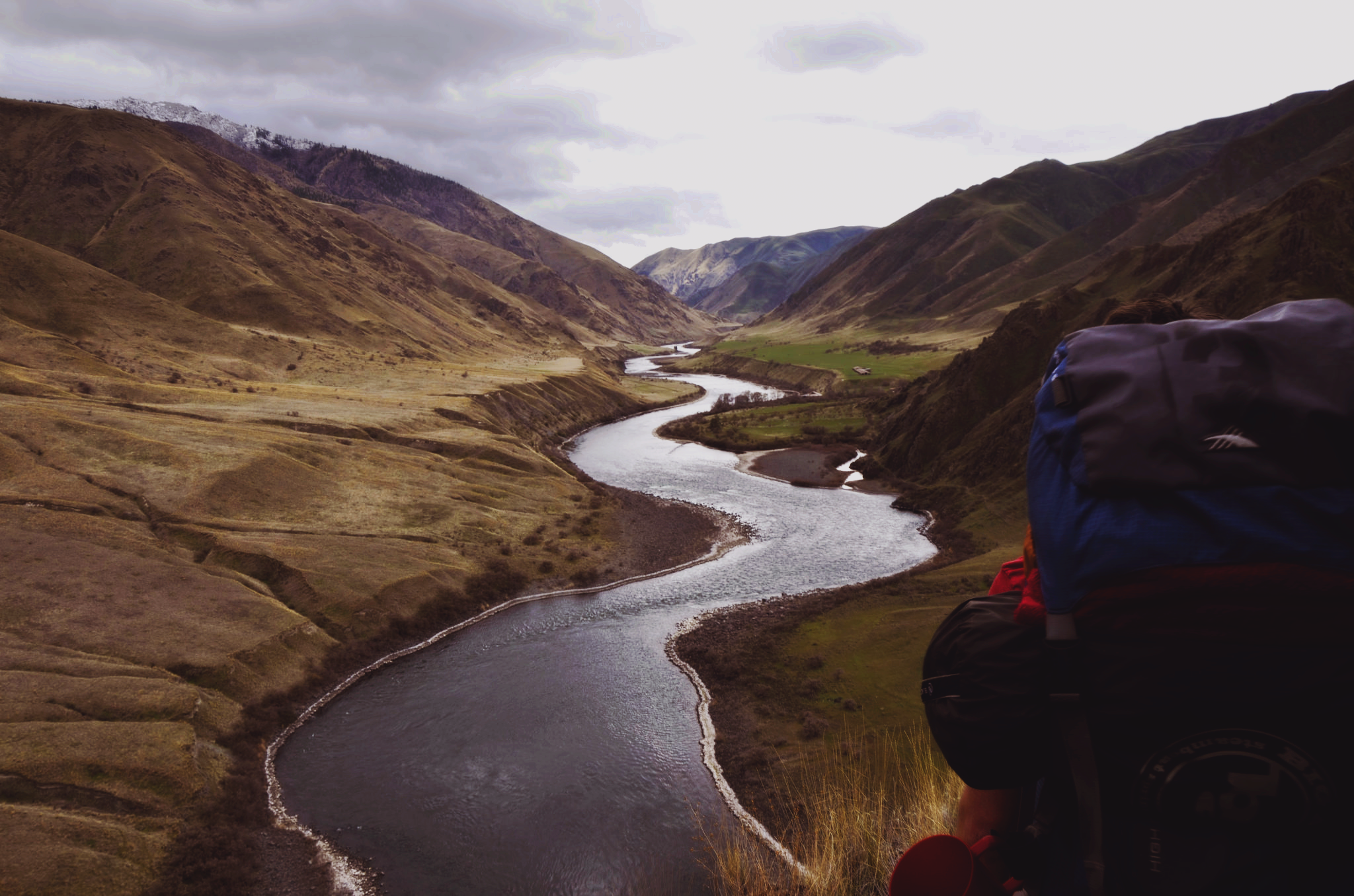 A winding river flows through a valley surrounded by grassy hills and mountains under a cloudy sky. A hiker with a large backpack is in the foreground, overlooking the scenic landscape.
