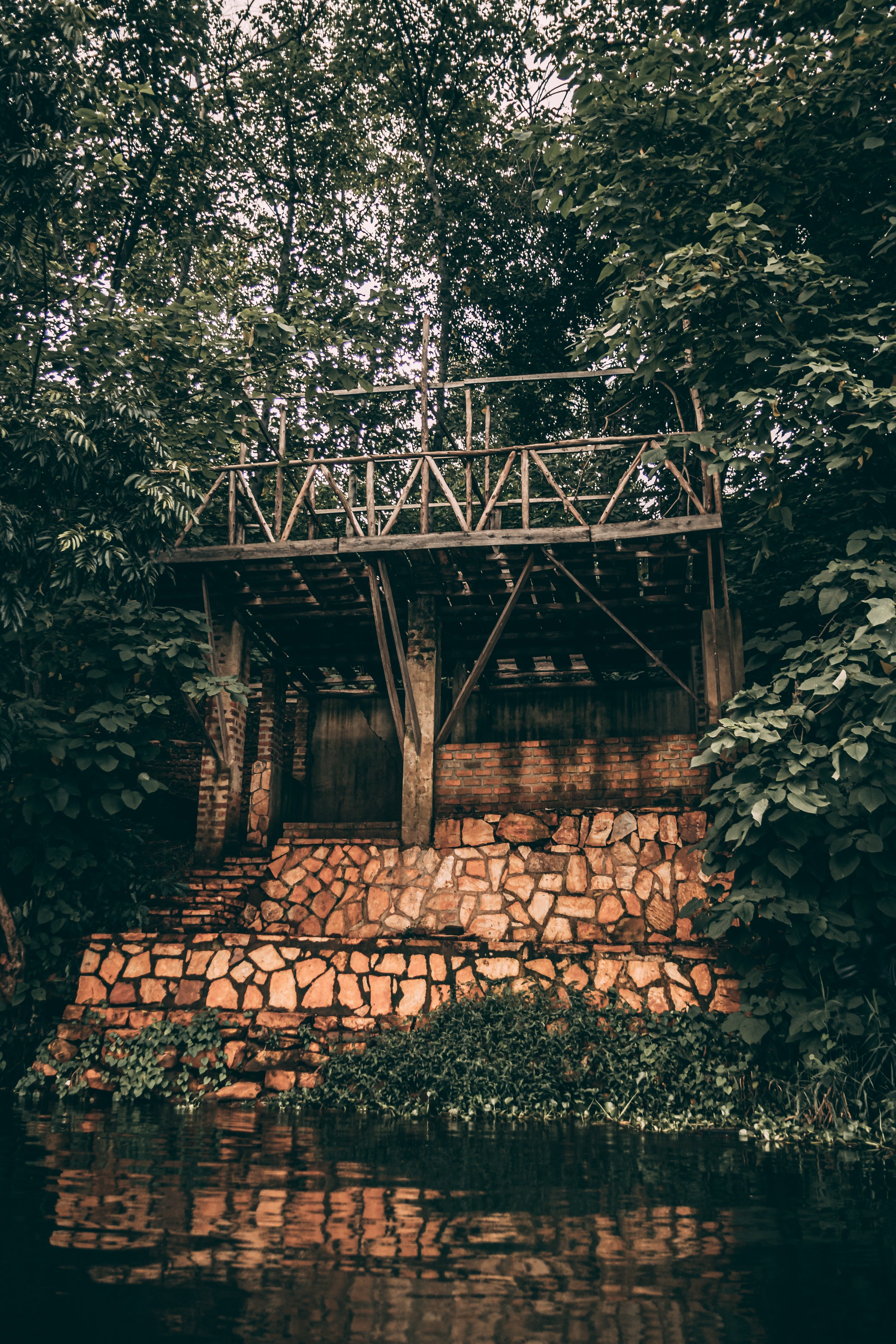 Wooden structure on stone platform surrounded by lush green trees beside a body of water.